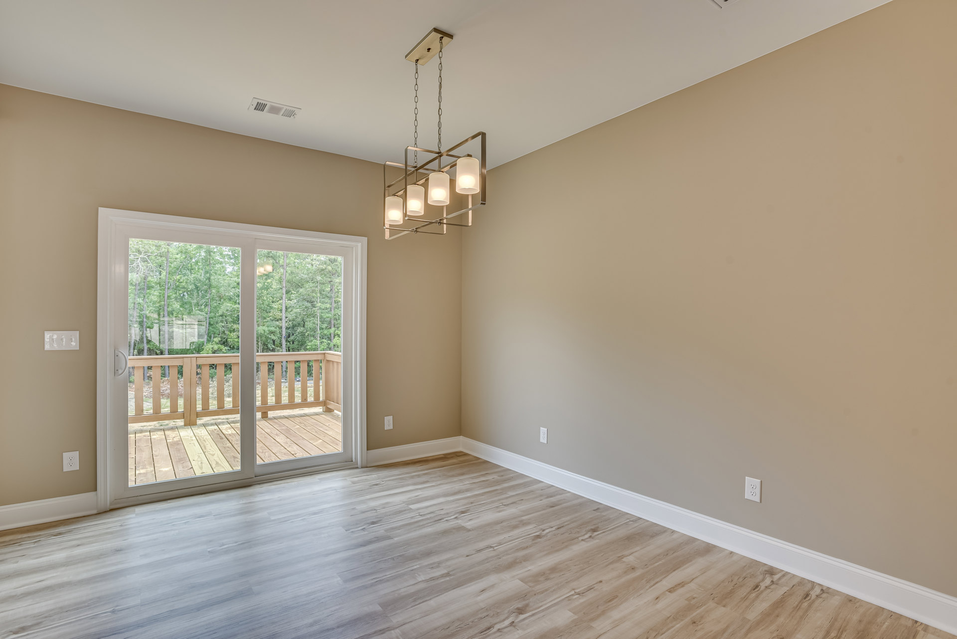 Wood flooring with white baseboards, sliding glass door opening to deck and trees, square-framed chandelier, wooden railing and post