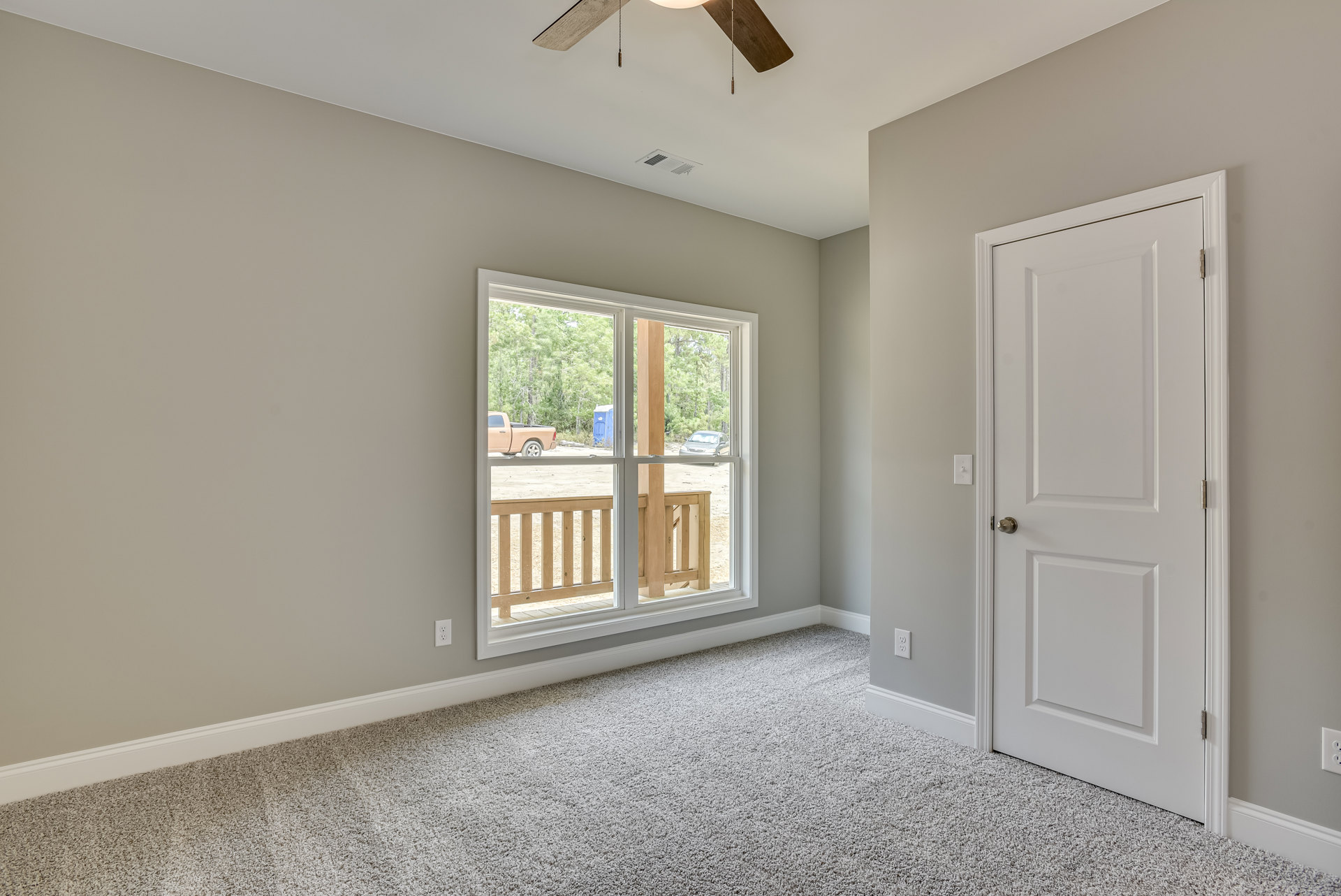 Carpeted bedroom with white walls, ceiling fan, large window overlooking wooden railing and parked truck, white door with silver knob