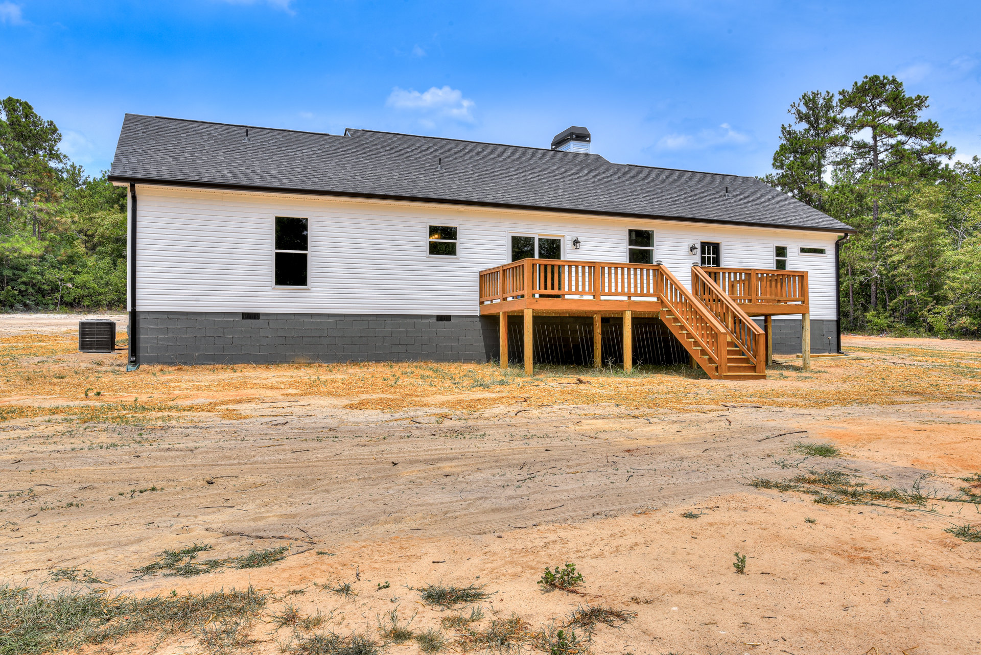 White house with wooden deck overlooking dirt road, surrounded by grassy land and trees under cloudy sky