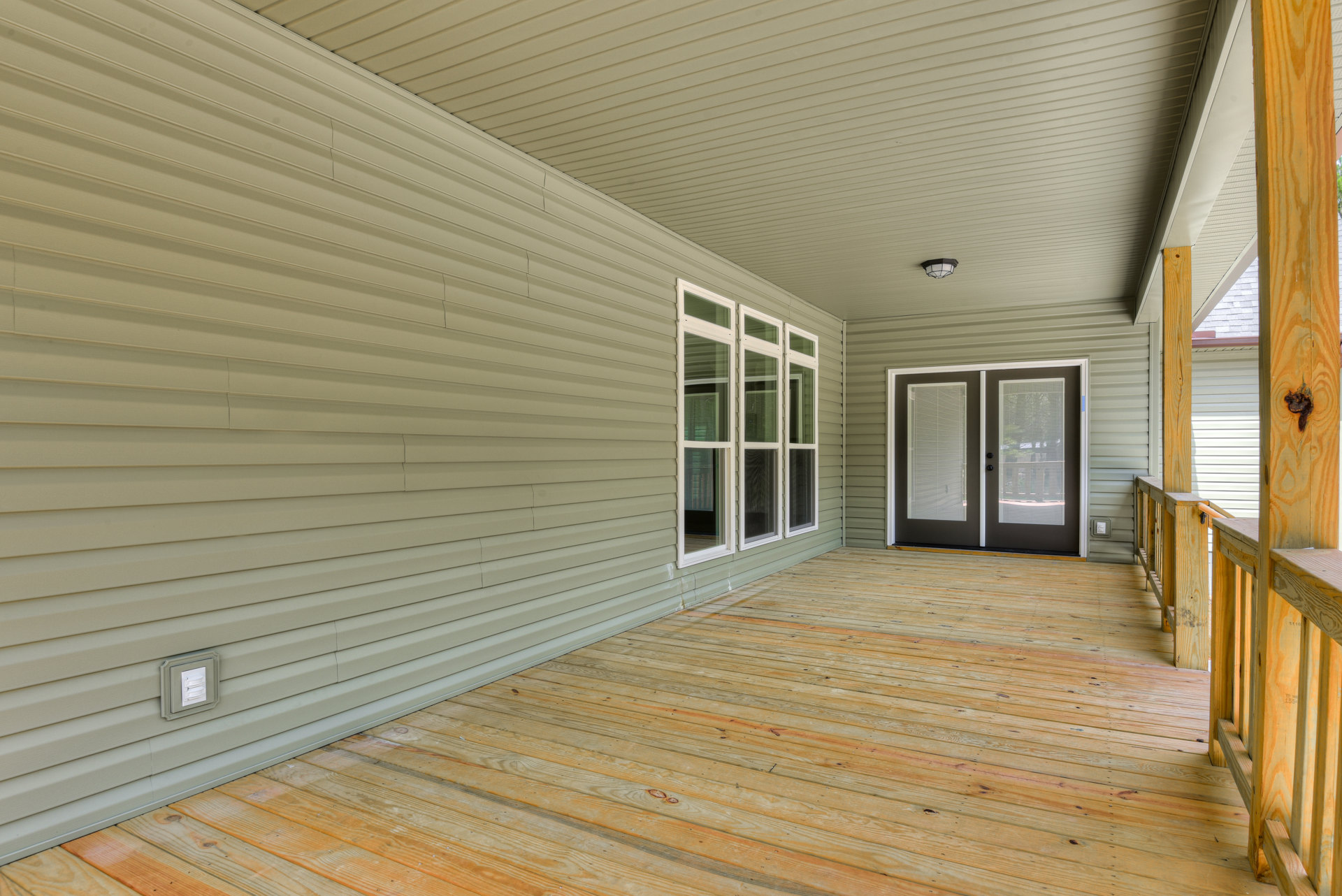 Covered porch with natural wood plank flooring, double doors with built-in blinds, white trim, and adjacent windows on a modern home exterior