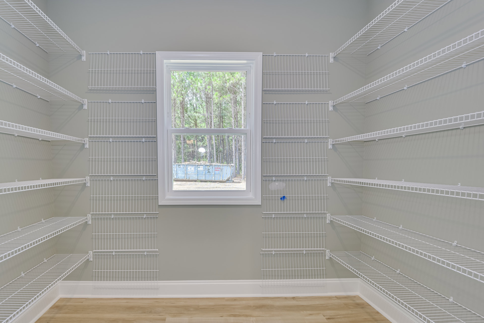 White wire shelving mounted on plaster walls, wood flooring, large window with outdoor view of trees and blue dumpster.