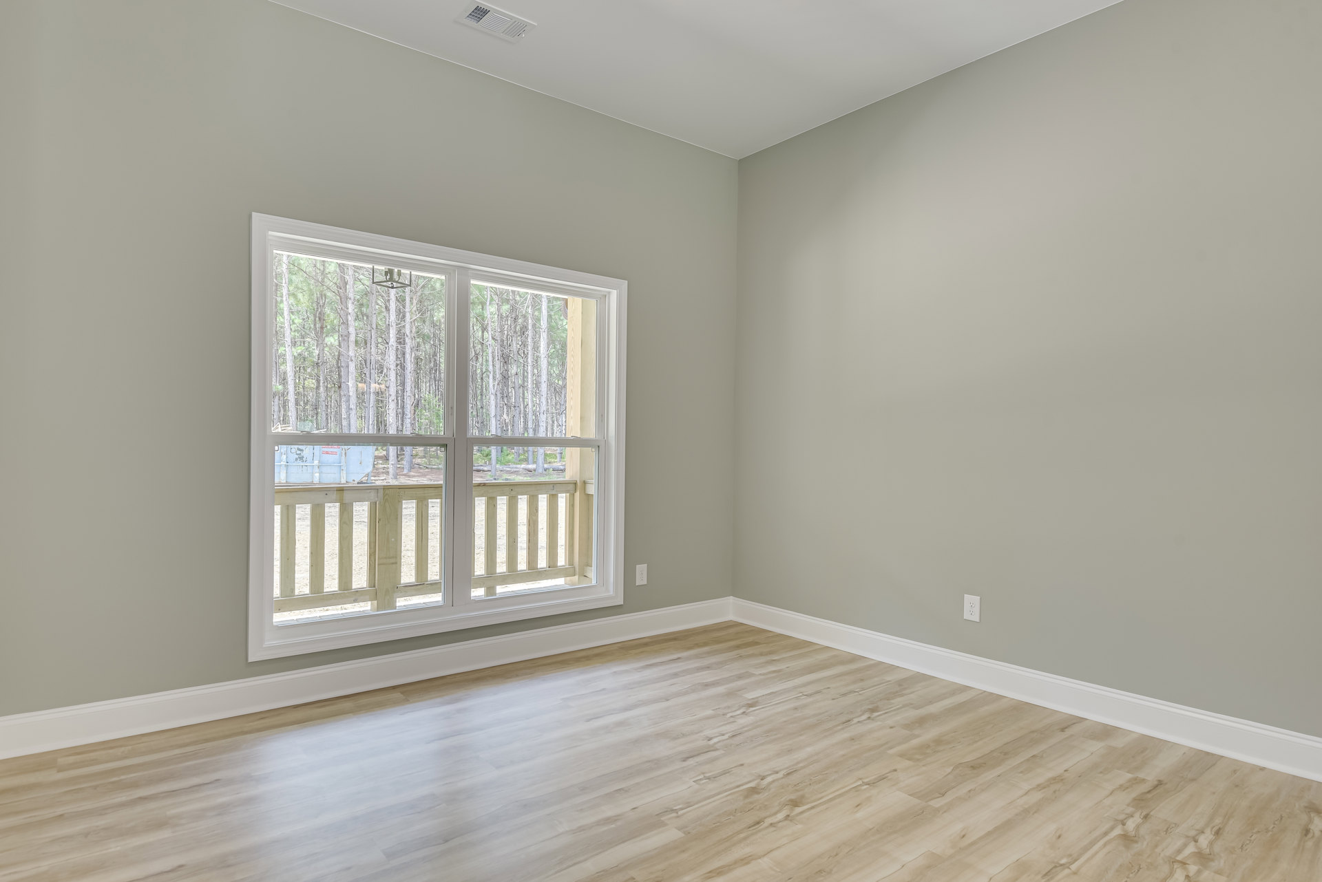 Room with white walls, wood flooring, large window framed by white trim, view of trees, porch, fence, and trash can outside, wooden railing visible through window