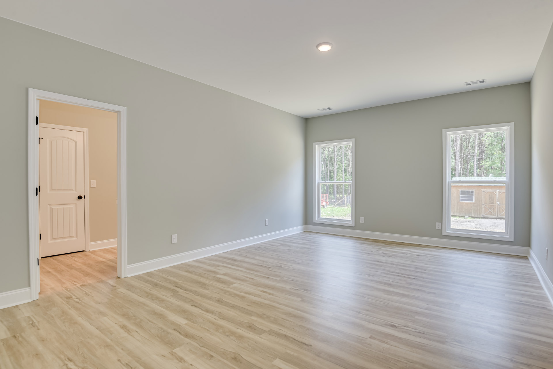 Spacious room with light wood flooring, large windows overlooking green trees, white walls, and a white door featuring a black handle