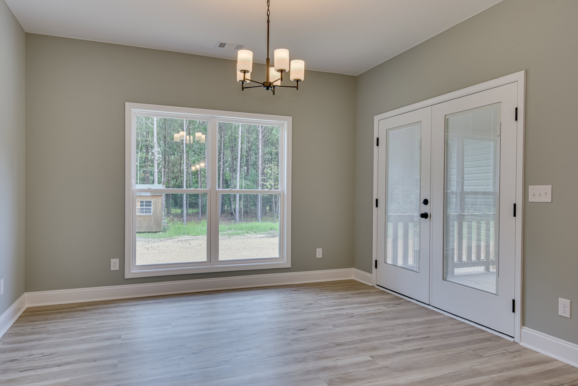 Hardwood floor room with white double doors featuring blinds, large windows showing trees outside, chandelier with white lamps, and a close-up lamp illuminating the space