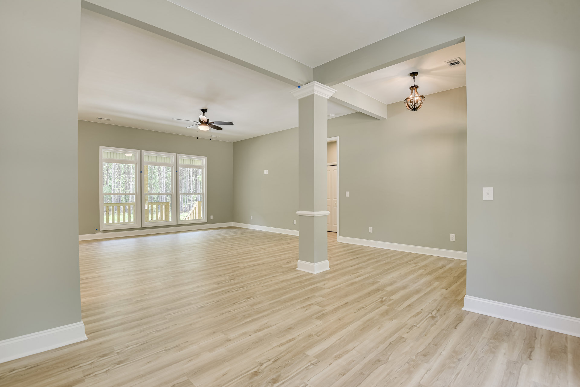 Room with wood flooring, white walls, ceiling fan with light, large window overlooking deck and trees.