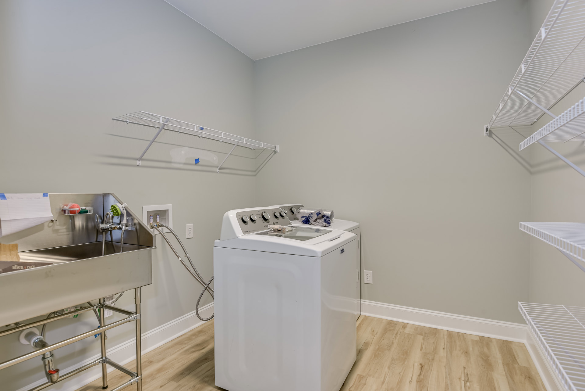 Front-loading washer and dryer set beside a white utility sink with chrome faucet, white wire shelving above, light cabinetry, and tile flooring in a laundry room.