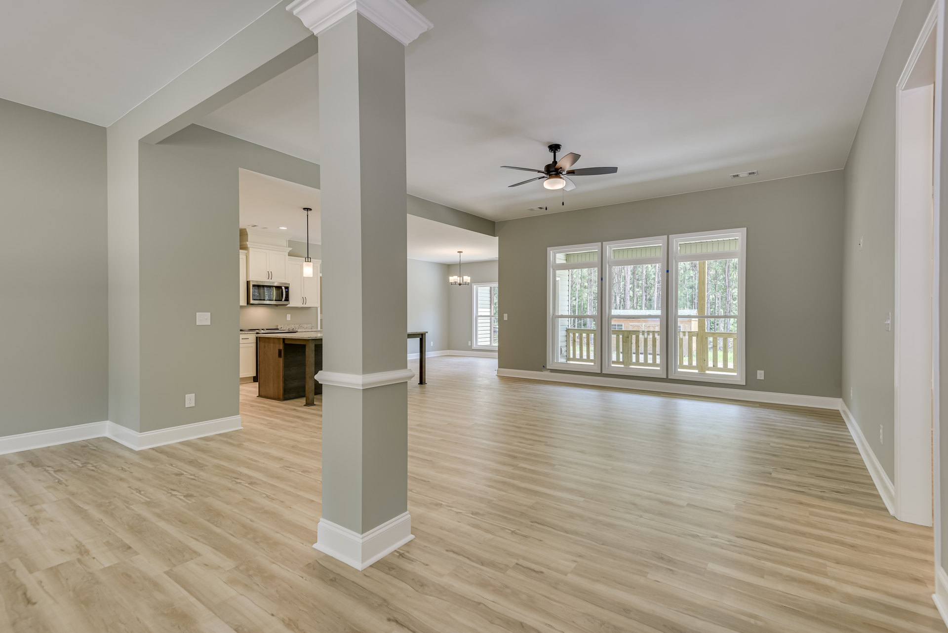 Spacious room featuring wood flooring, white trim, ceiling fan with light, window with wood railing overlooking trees, and kitchen island with sink