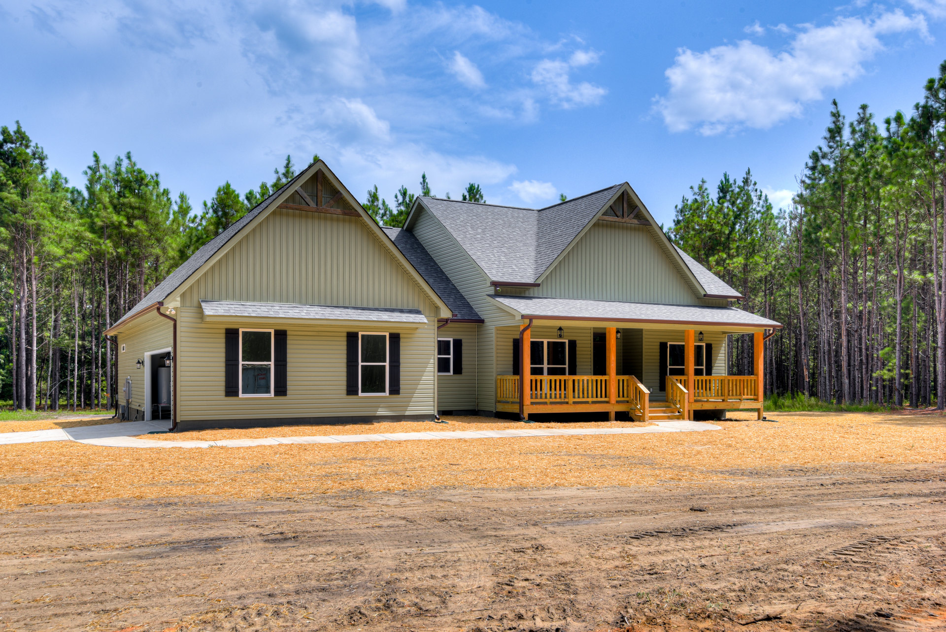 Two-story cottage-style home with black-shuttered windows, wooden porch railing, surrounded by mature trees, dirt driveway in foreground, gray roof partially visible against cloudy