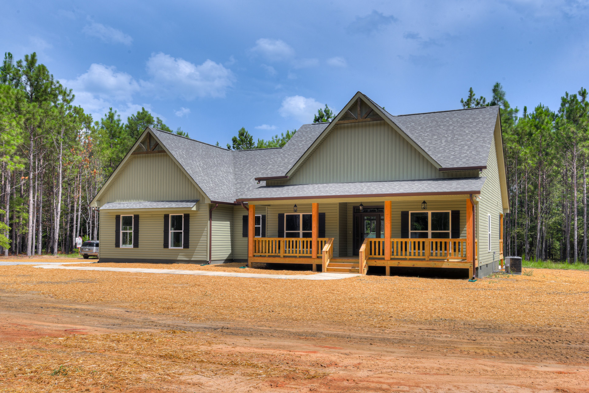 Two-story house with white-framed windows, wooden porch and bench, silver SUV parked on a dirt road, surrounded by trees under a cloudy sky