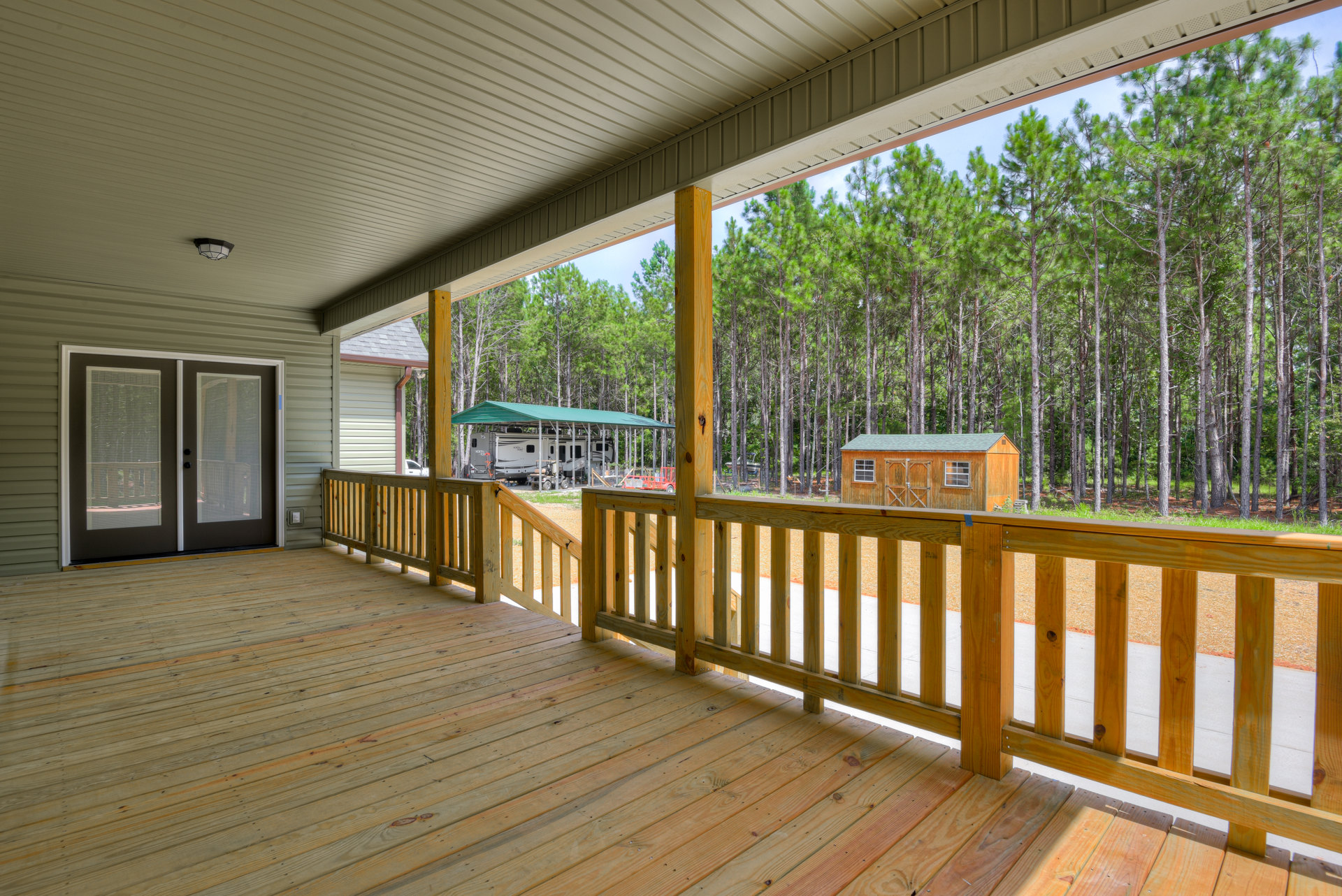 Wooden deck with railings and covered porch, double doors with blinds, view of trees, neighboring house, RV parked under canopy, wooden shed with window and door