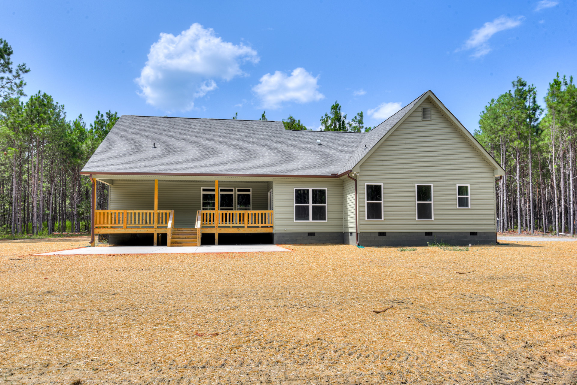 Large covered porch with white railings, wide steps, and wooden bench; grey siding, white-framed windows, attached garage with paneled door, surrounded by trees and cloudy sky
