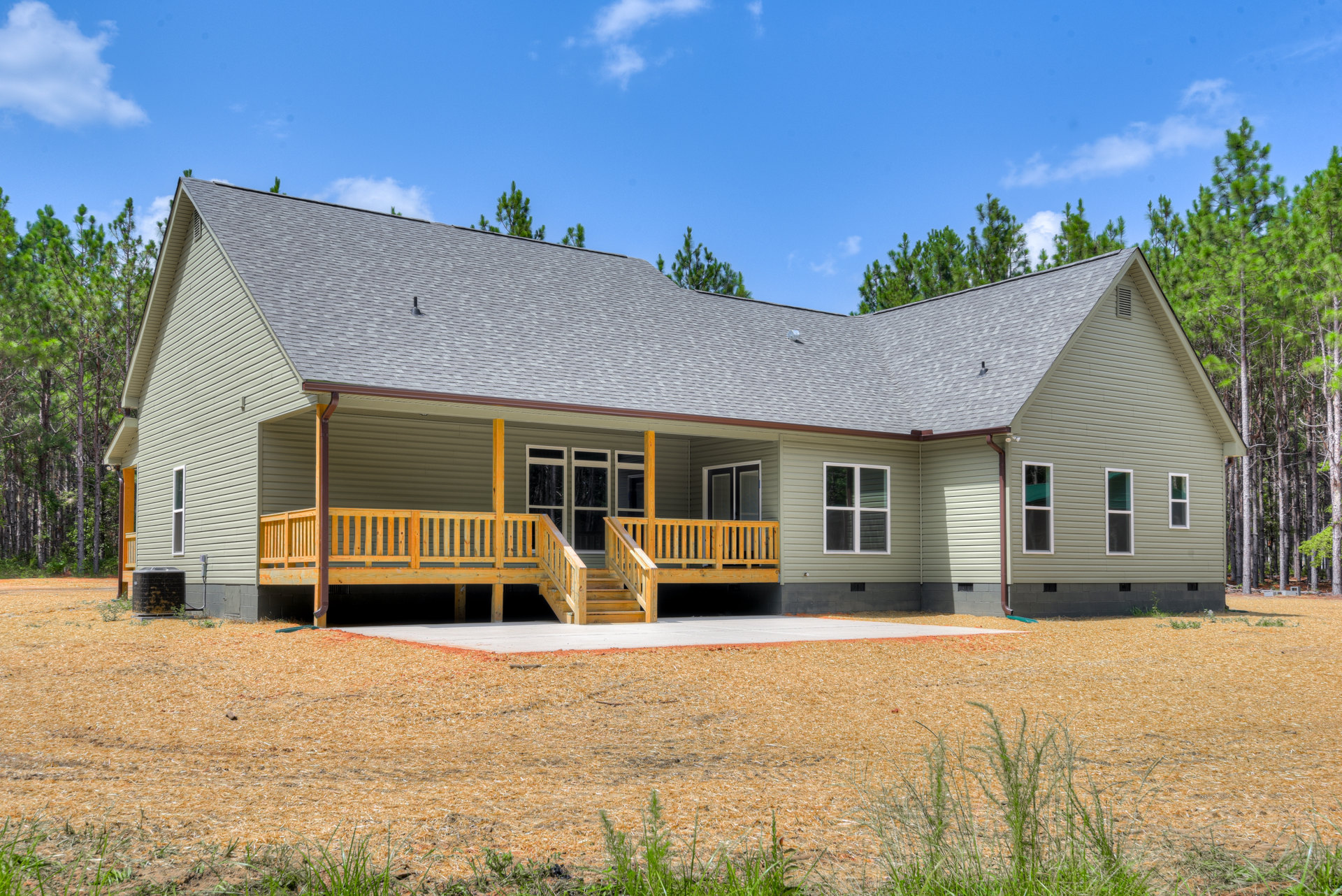 Two-story house with white-framed windows, wooden porch and deck, wooden staircase and railing, concrete driveway, surrounded by trees and landscaping under a partly cloudy sky