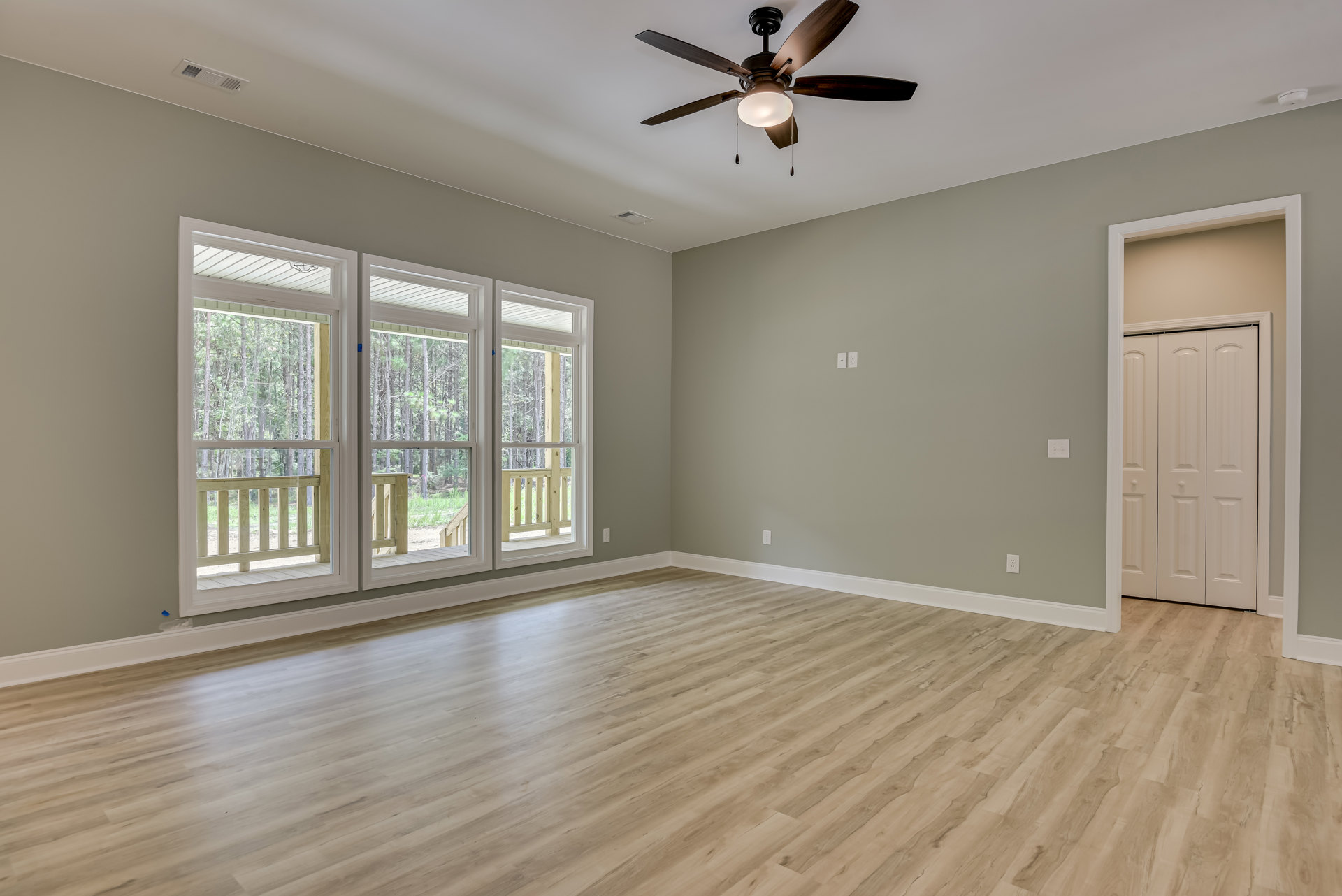 Ceiling fan with light fixture, hardwood flooring, white closet doors with round knobs, windows overlooking deck and trees, white walls with crown molding