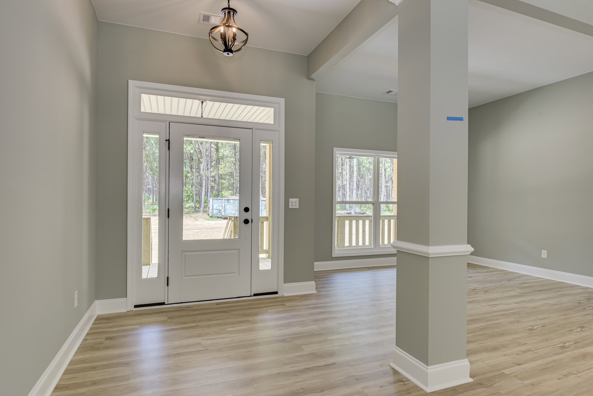 White-paneled door with glass panes, wood flooring with white baseboard trim, ceiling light fixture, window overlooking green trees, white utility box visible outside in grass