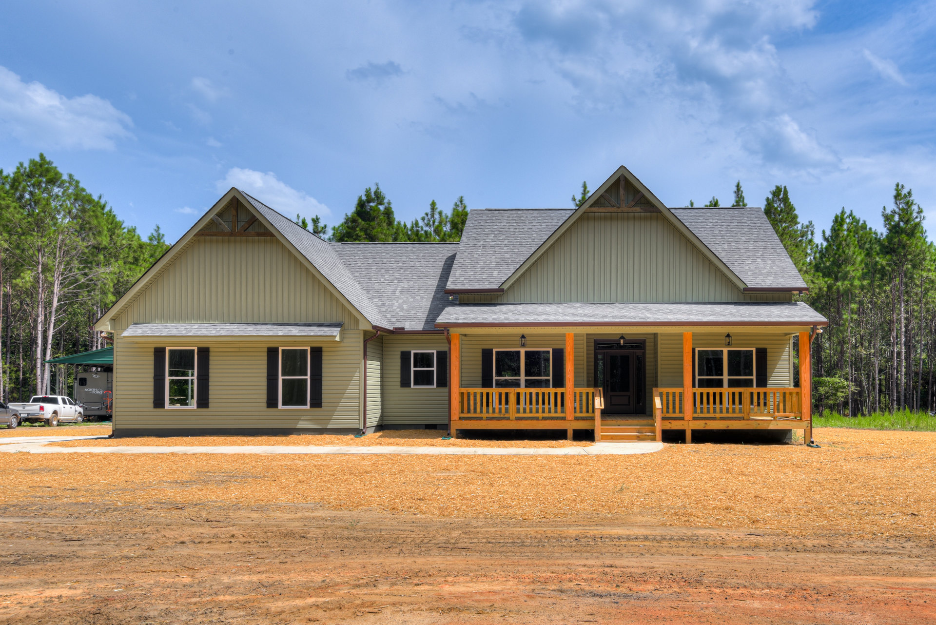 Two-story house with covered porch, black shuttered windows, gray roof, surrounded by dirt, wood chips, and mature trees; white truck parked on grassy area.