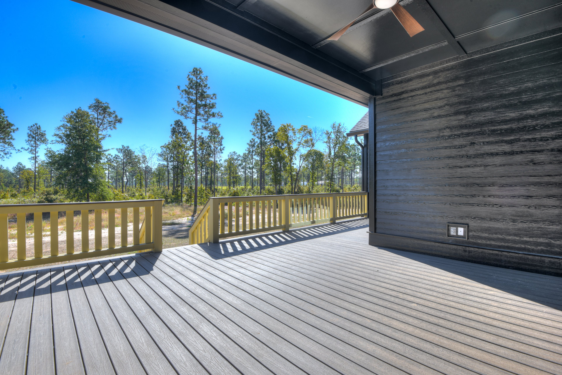 Wood deck with white and yellow railings, ceiling fan overhead, surrounded by leafy trees and blue sky in the background