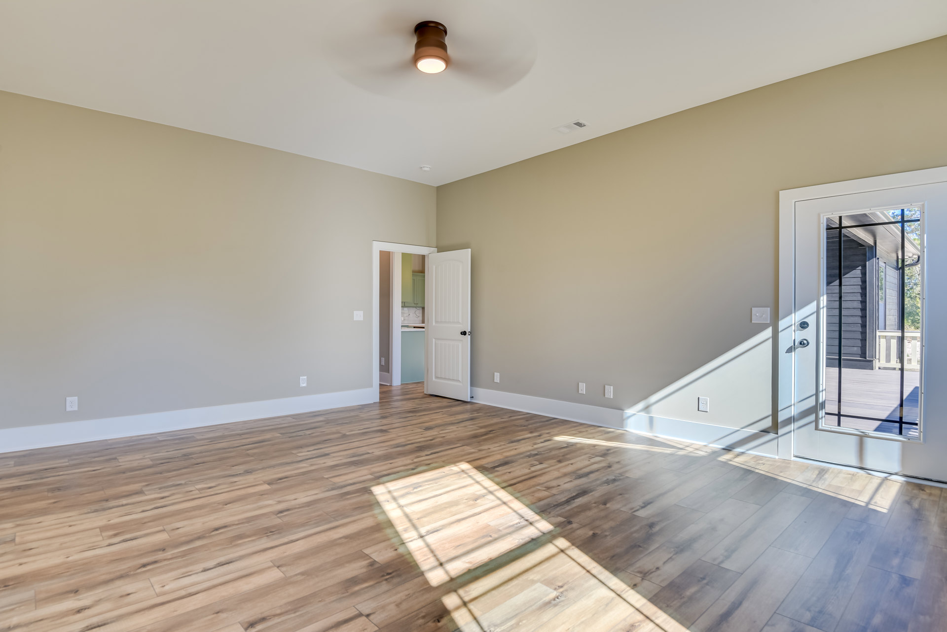 Wood flooring in a bright room with a ceiling fan, white walls, glass patio door, and a white interior door with black hardware.