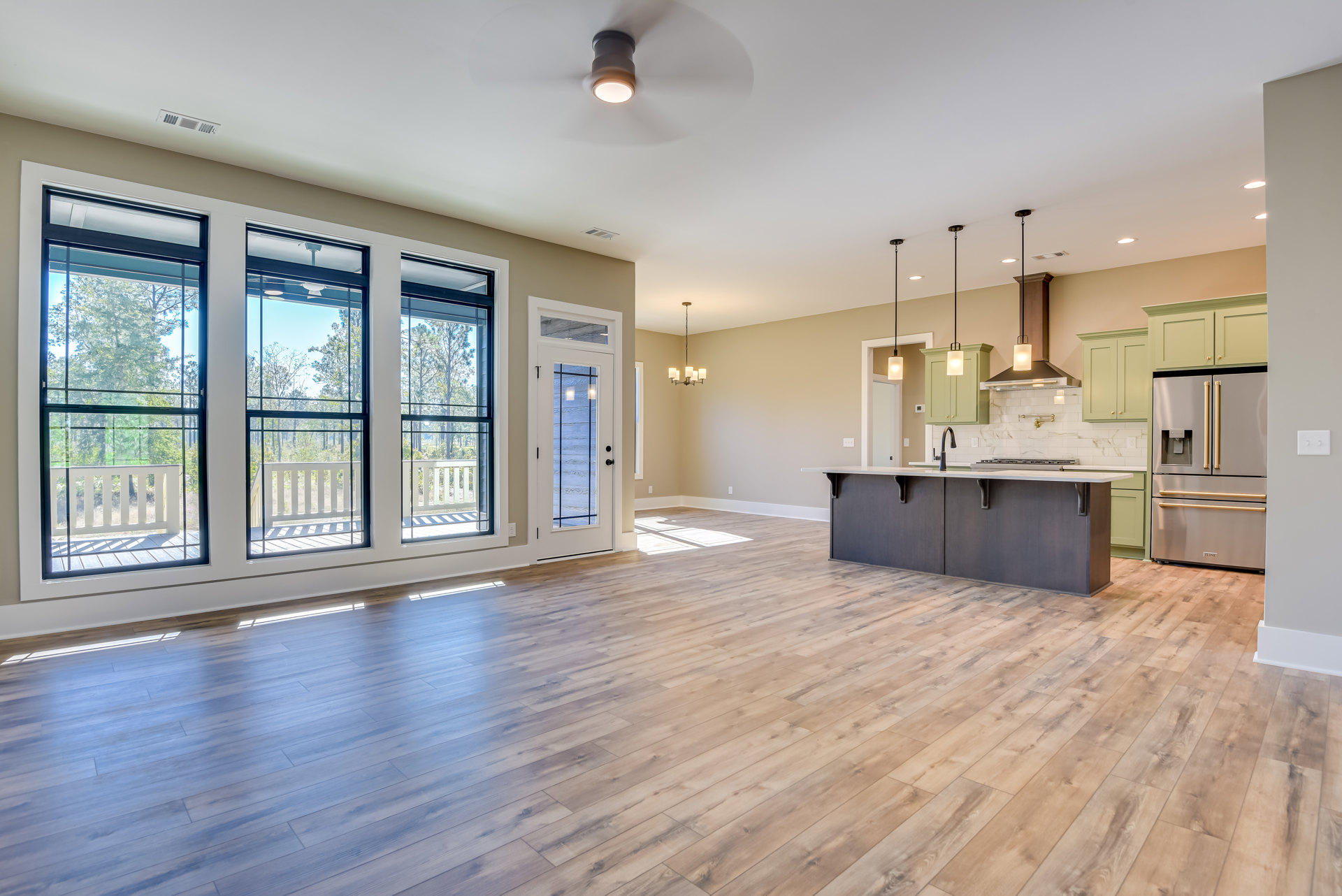 Open kitchen with wood flooring, central island, stainless refrigerator with water dispenser, white cabinetry, sink and stove on kitchen counter, large windows overlooking trees