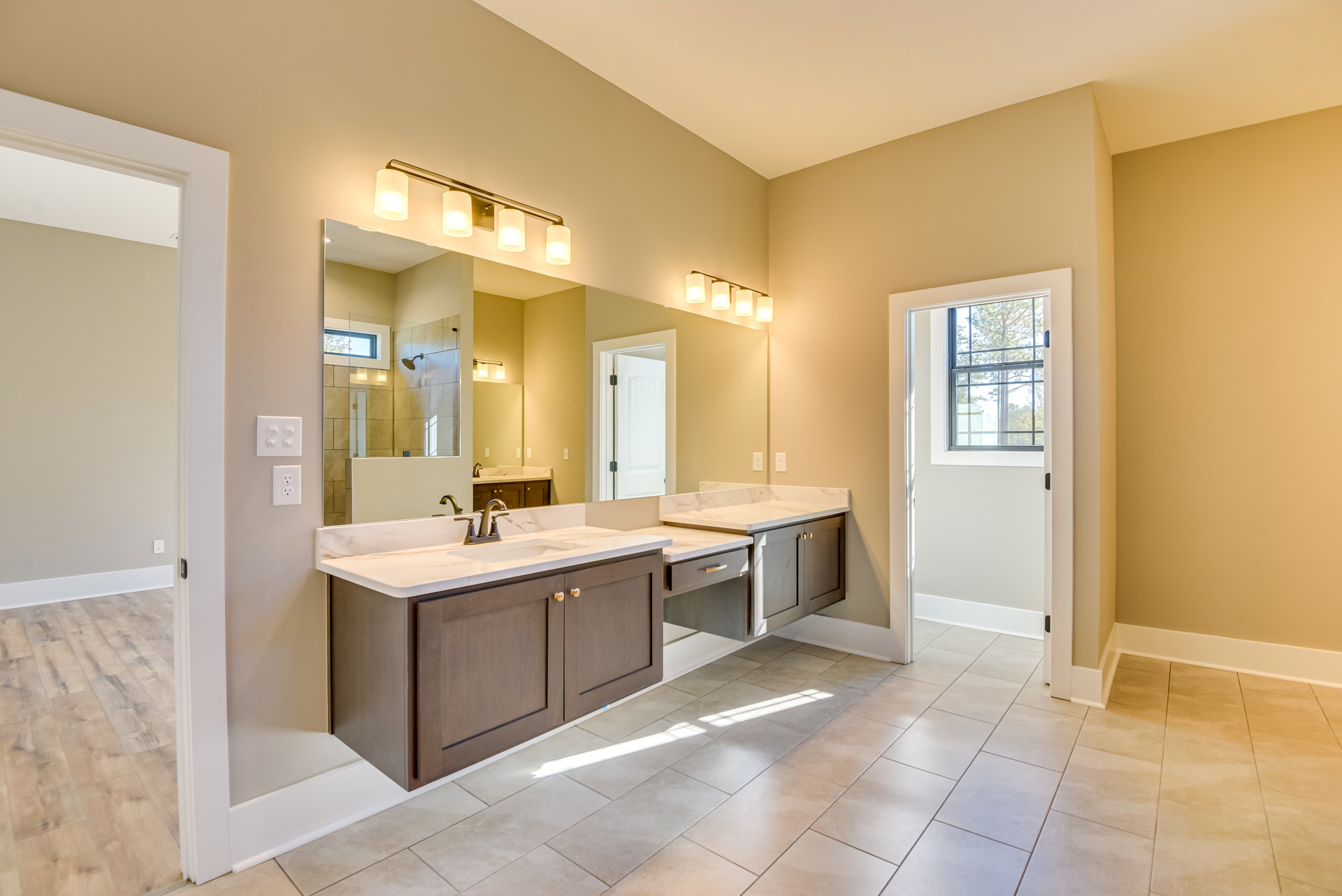 Bathroom with two rectangular sinks set in a stone countertop, large frameless mirror above, white cabinetry, black hardware, tile flooring, window showing leafy tree outside.