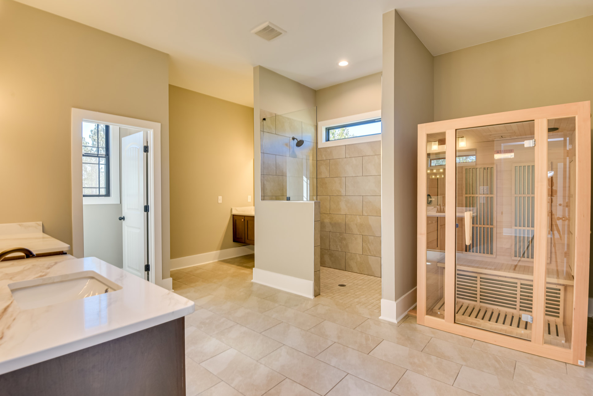 Modern bathroom featuring a glass shower enclosure, white countertop with integrated sink, light tile flooring, white walls, and a window beside a white door.
