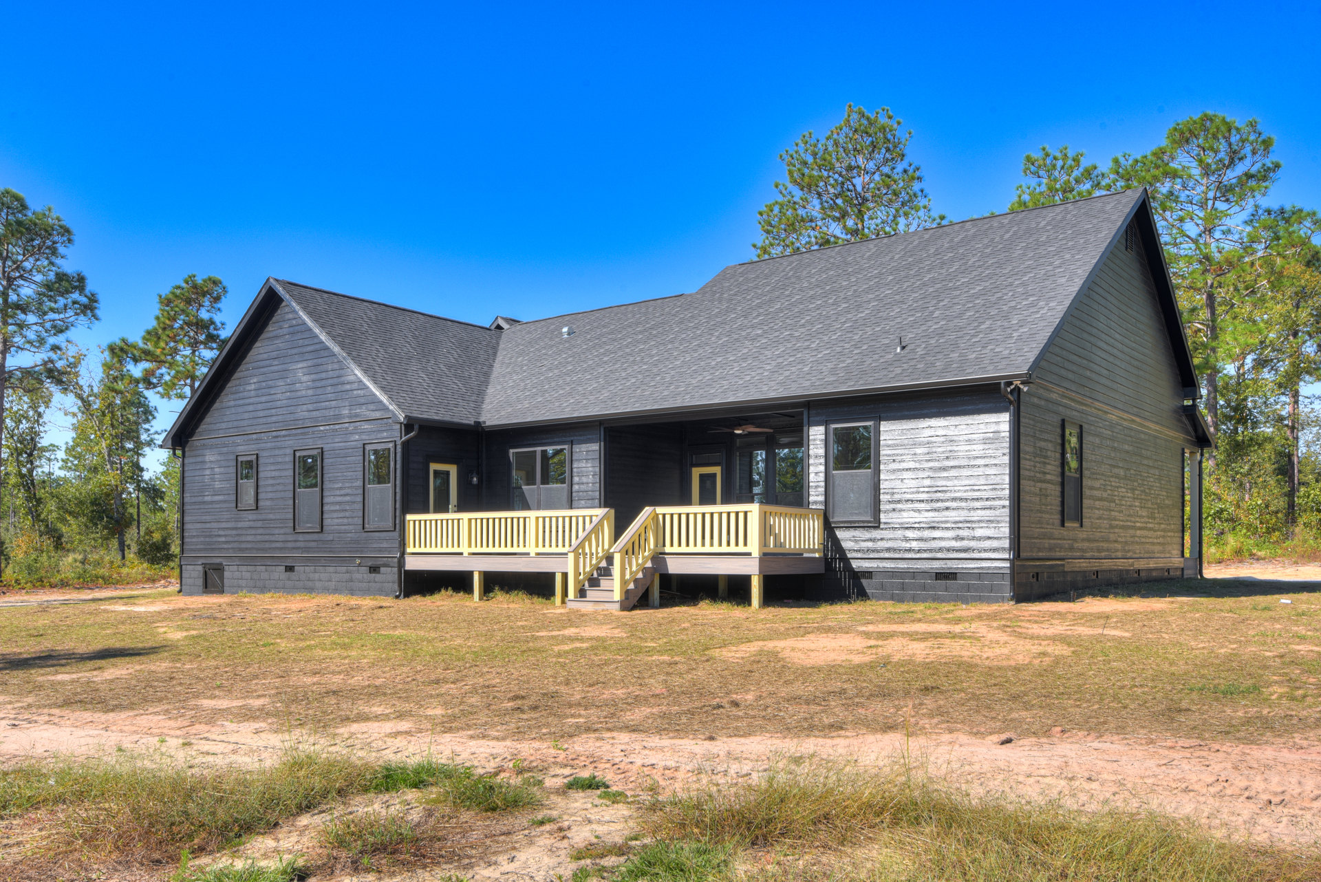 Wooden porch with yellow railings, wooden staircase leading to grassy yard, cottage-style house with large window reflecting trees, sloped roof with tree in background.