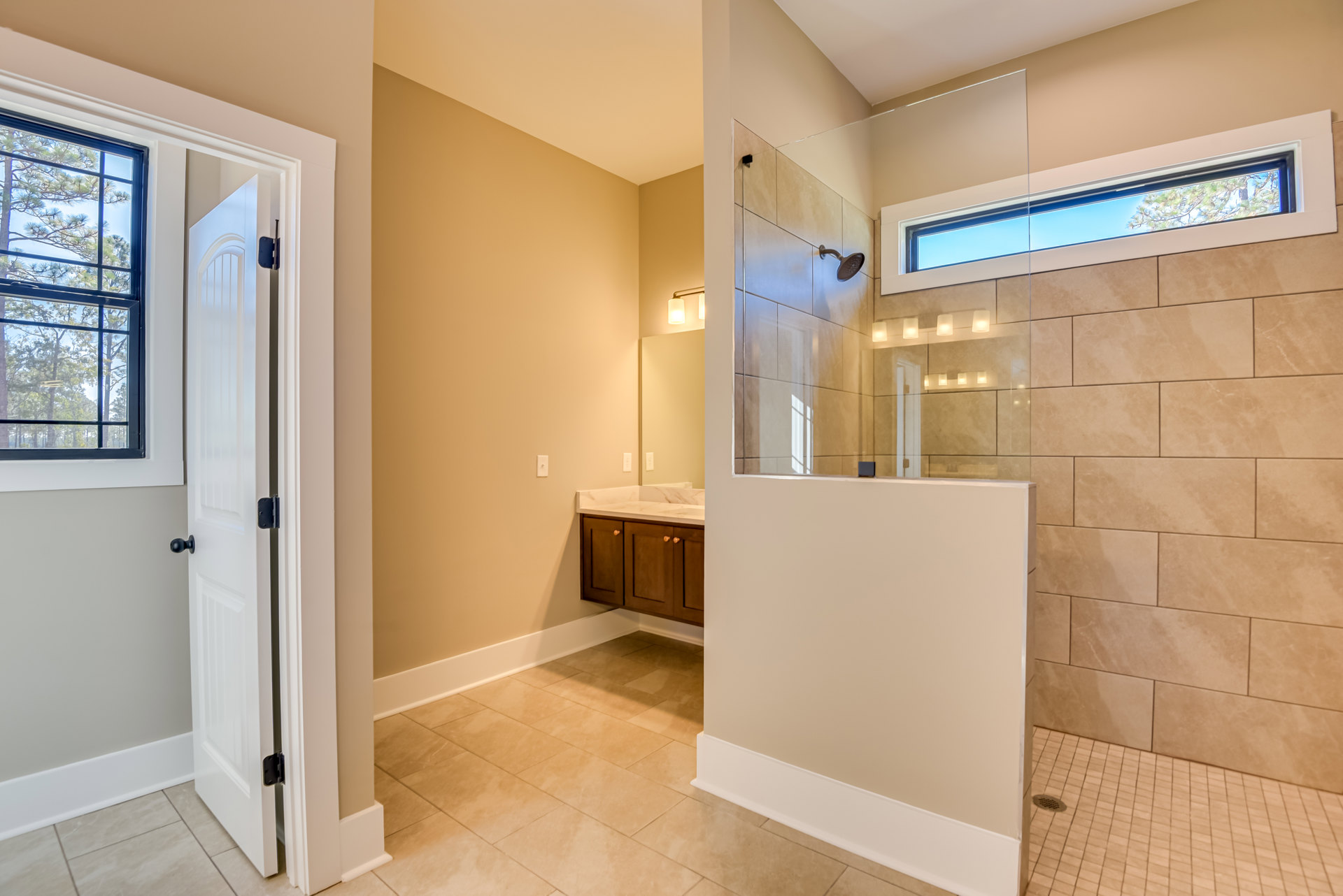 Bathroom with glass shower enclosure, white sink, white walls and baseboards, window overlooking trees, white door with black knob