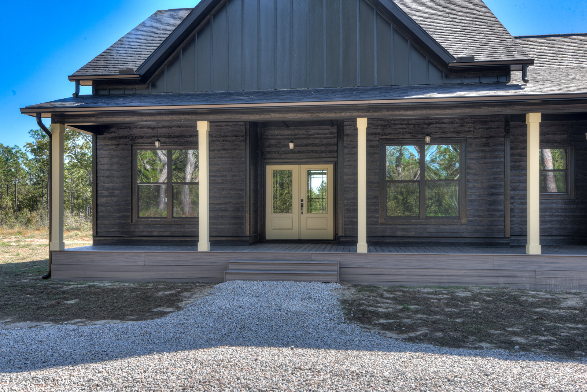 Front porch with double glass doors, white siding, gravel pile beside wall, large window reflecting trees, metal accent wall, gabled roof, landscaped yard.