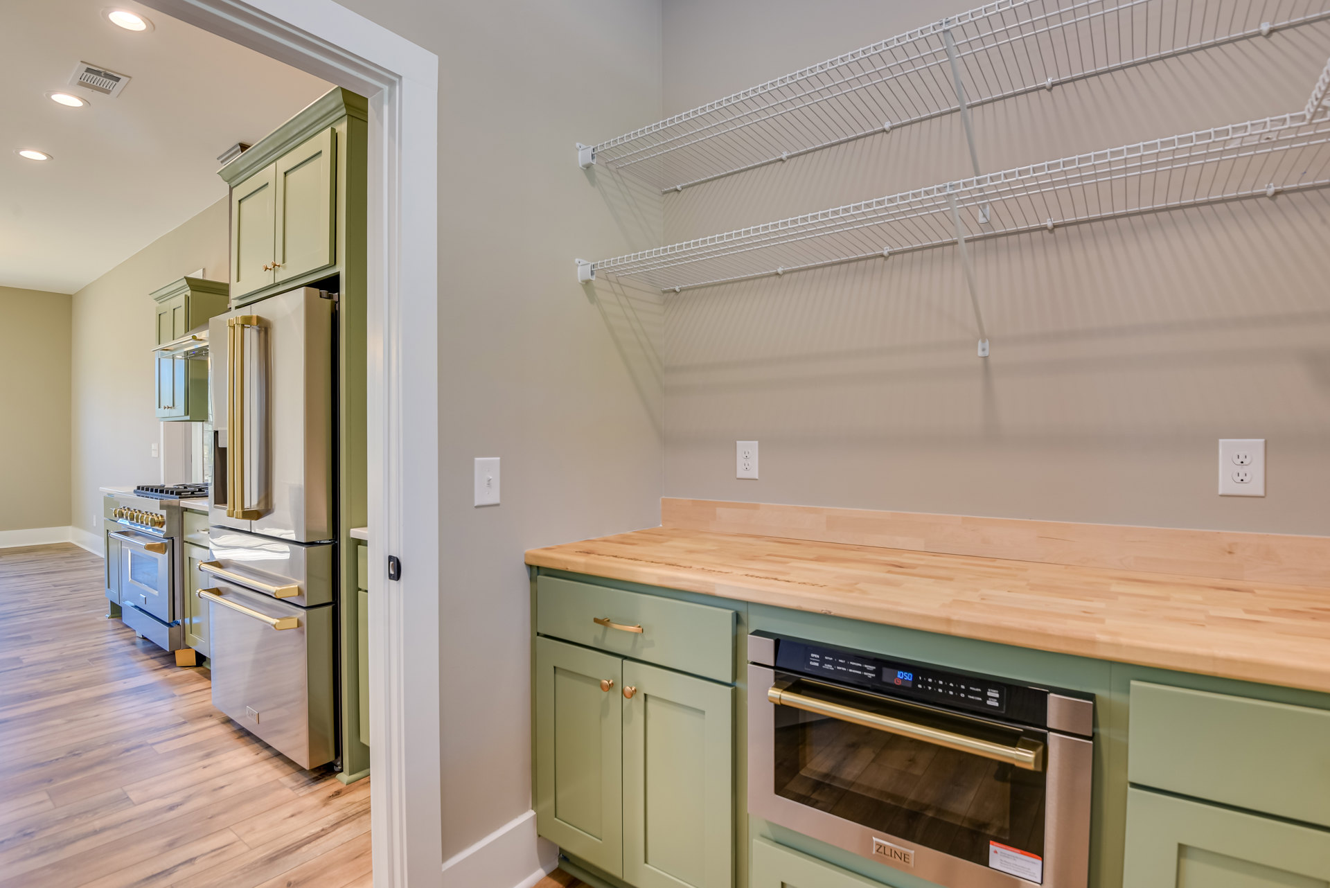 Kitchen with green cabinets, wood countertop, stainless steel refrigerator with gold handle, white wire shelving on wall, close-up of oven, outlet, and countertop