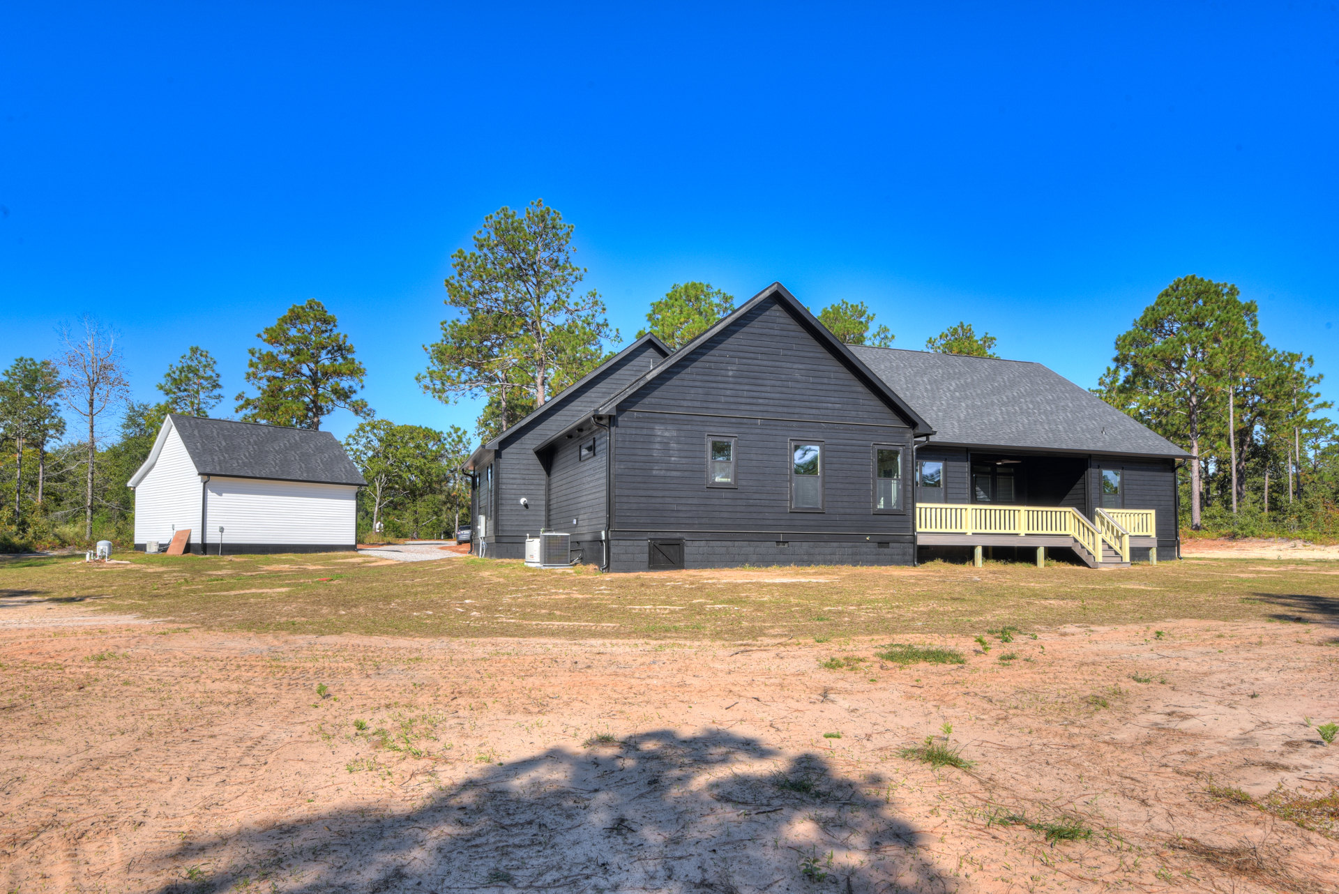 White house with black roof, attached garage, dirt yard, deck with yellow railing, tree shadows on ground, trees in background
