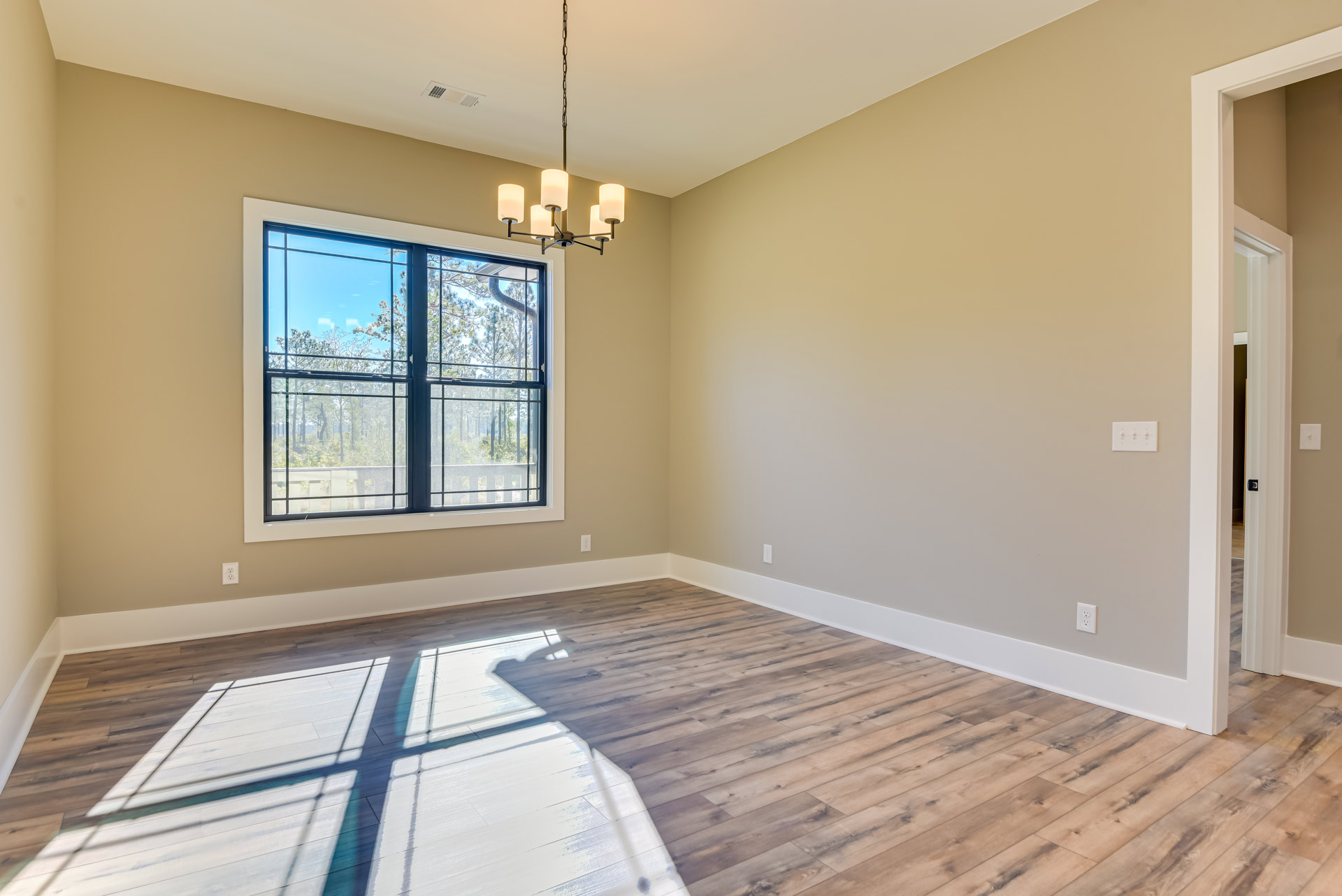 Sunlit room with wide wood plank flooring, white plaster walls, large window overlooking trees, and a modern chandelier hanging from the ceiling