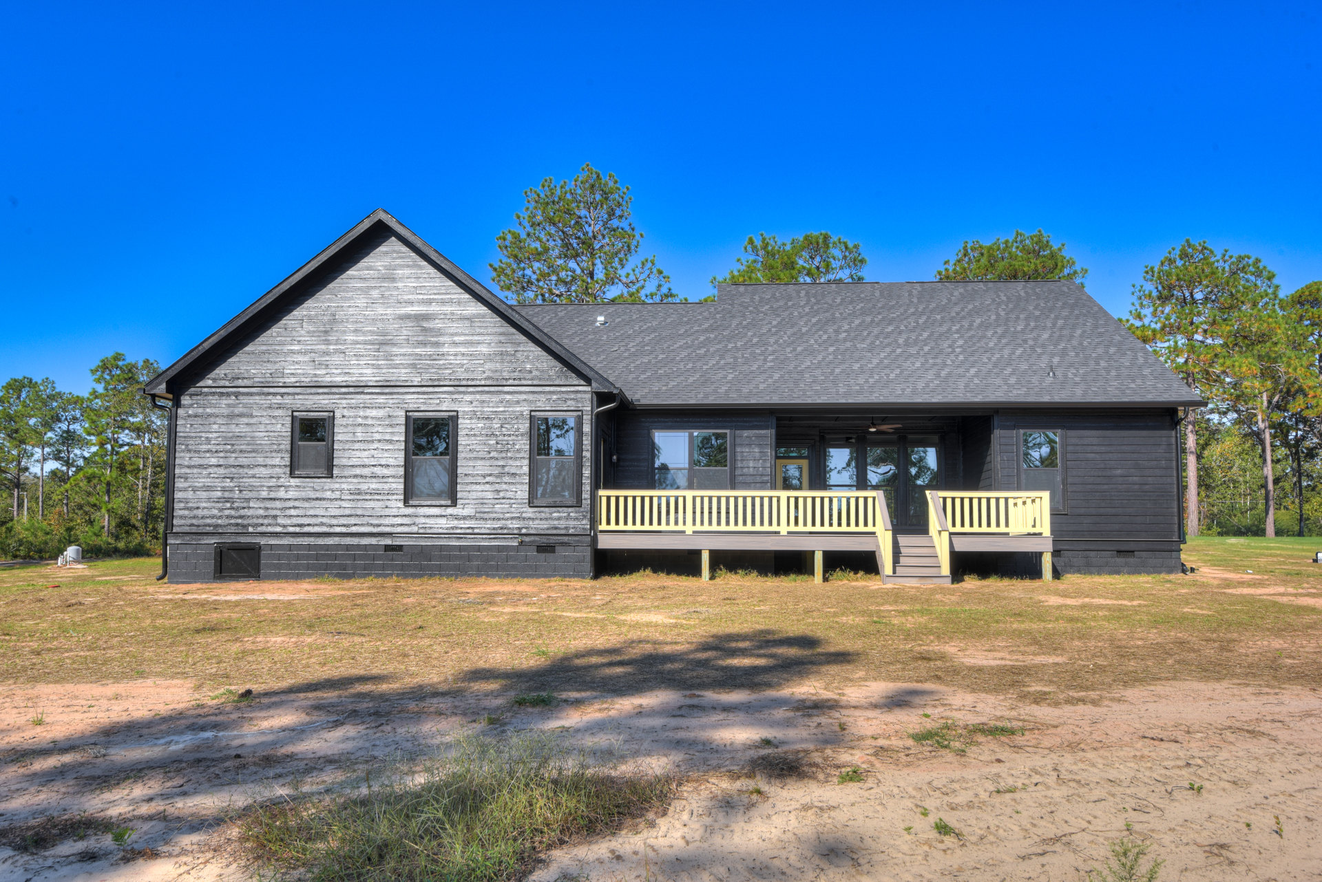 Two-story cottage with yellow porch railing, glass windows reflecting trees, gray roof, and grassy yard.