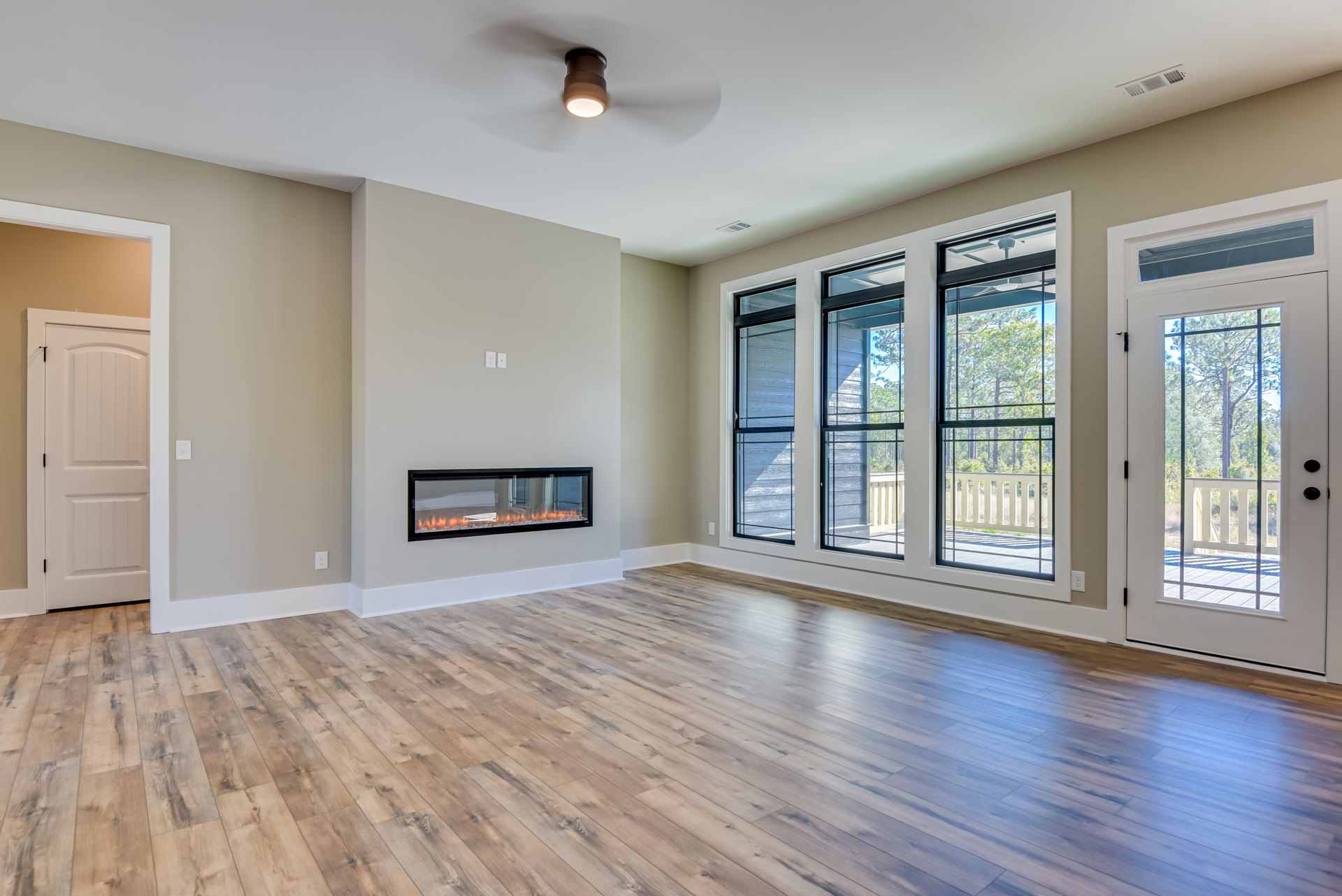 Living room with hardwood floor, modern fireplace set in glass wall, white door with black handle, large window overlooking trees and porch