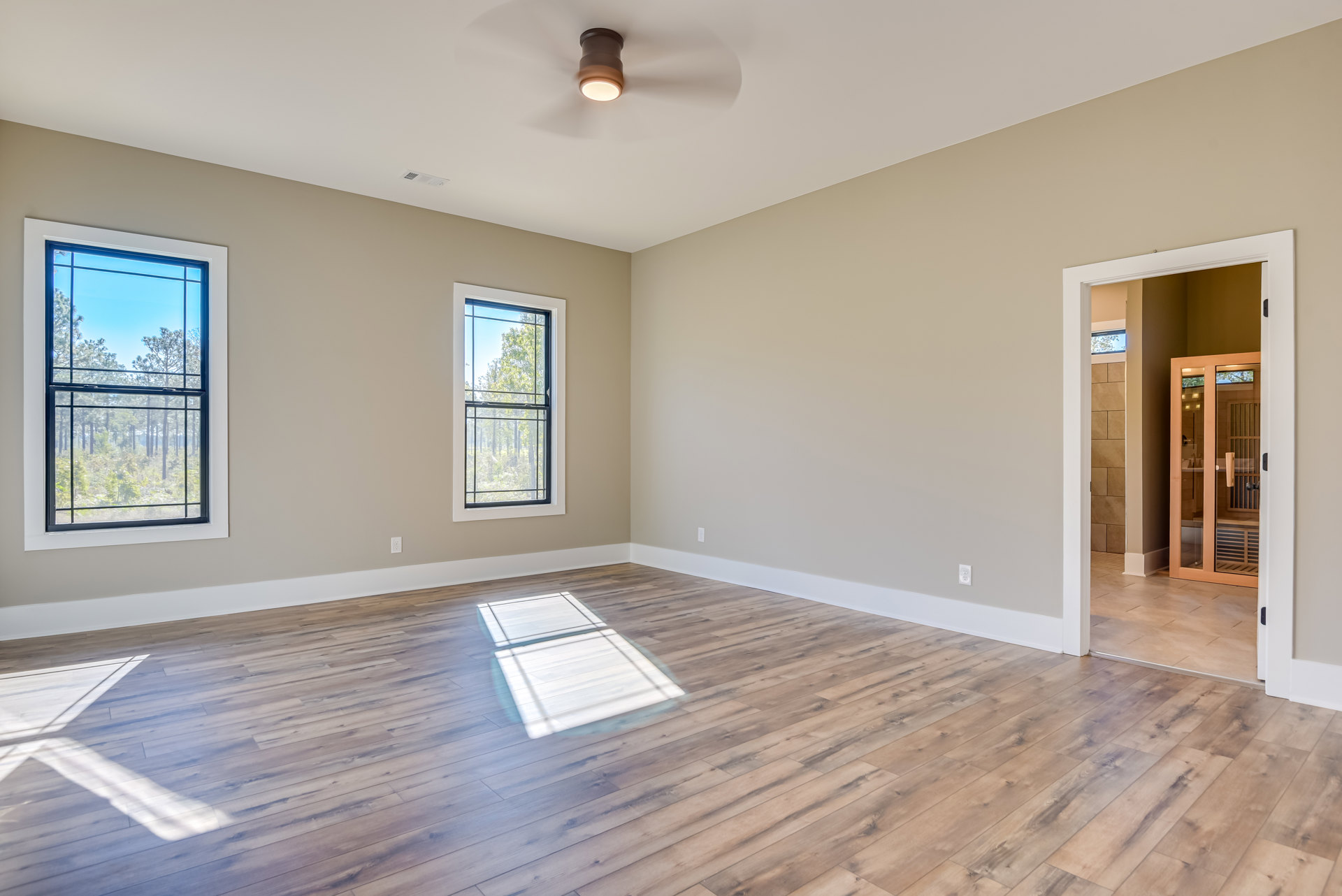 Wood flooring with white tile accent, ceiling fan overhead, glass door and large window revealing trees outside, sunlight illuminating the space