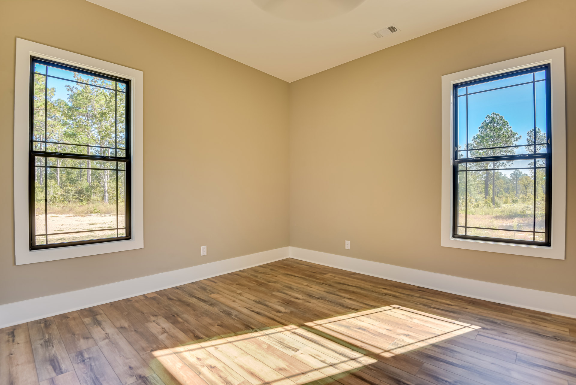 Sunlit room featuring wide plank wood flooring, large windows with views of leafy trees, and neutral walls.