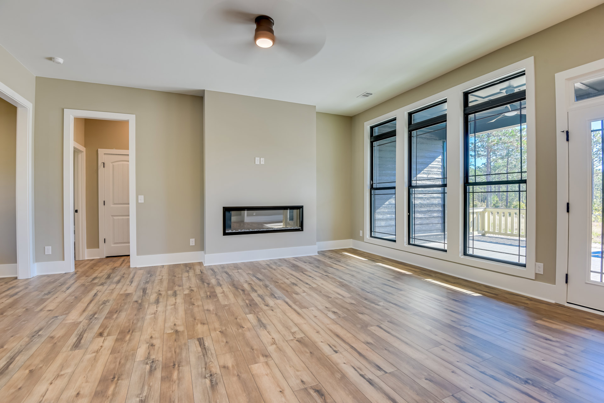 Living room with hardwood floors, white walls, black-framed fireplace, large window showing trees, modern light fixture, and white door with black handle