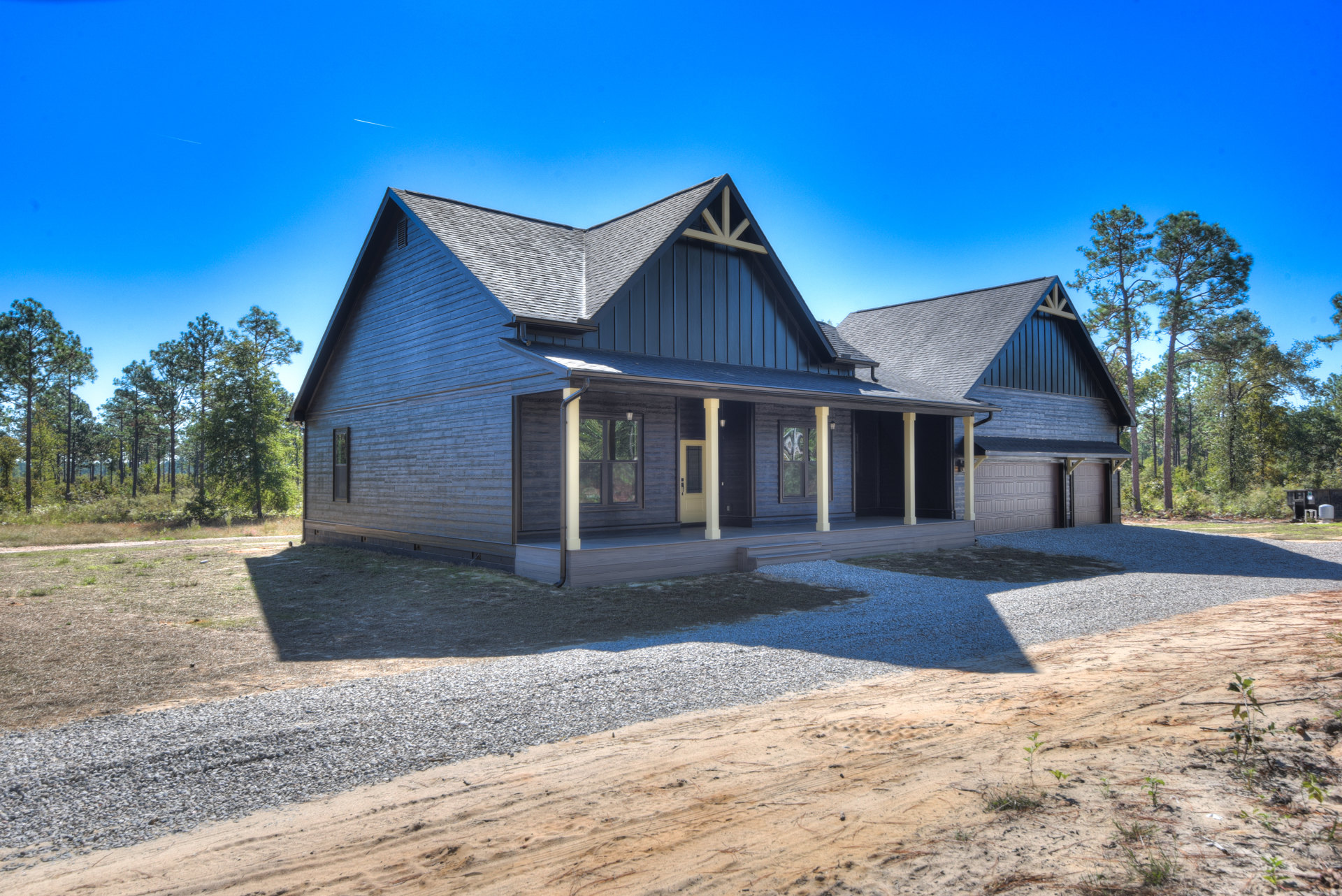 Two-story home with gray siding, covered front porch, gravel driveway, and mature trees lining the background