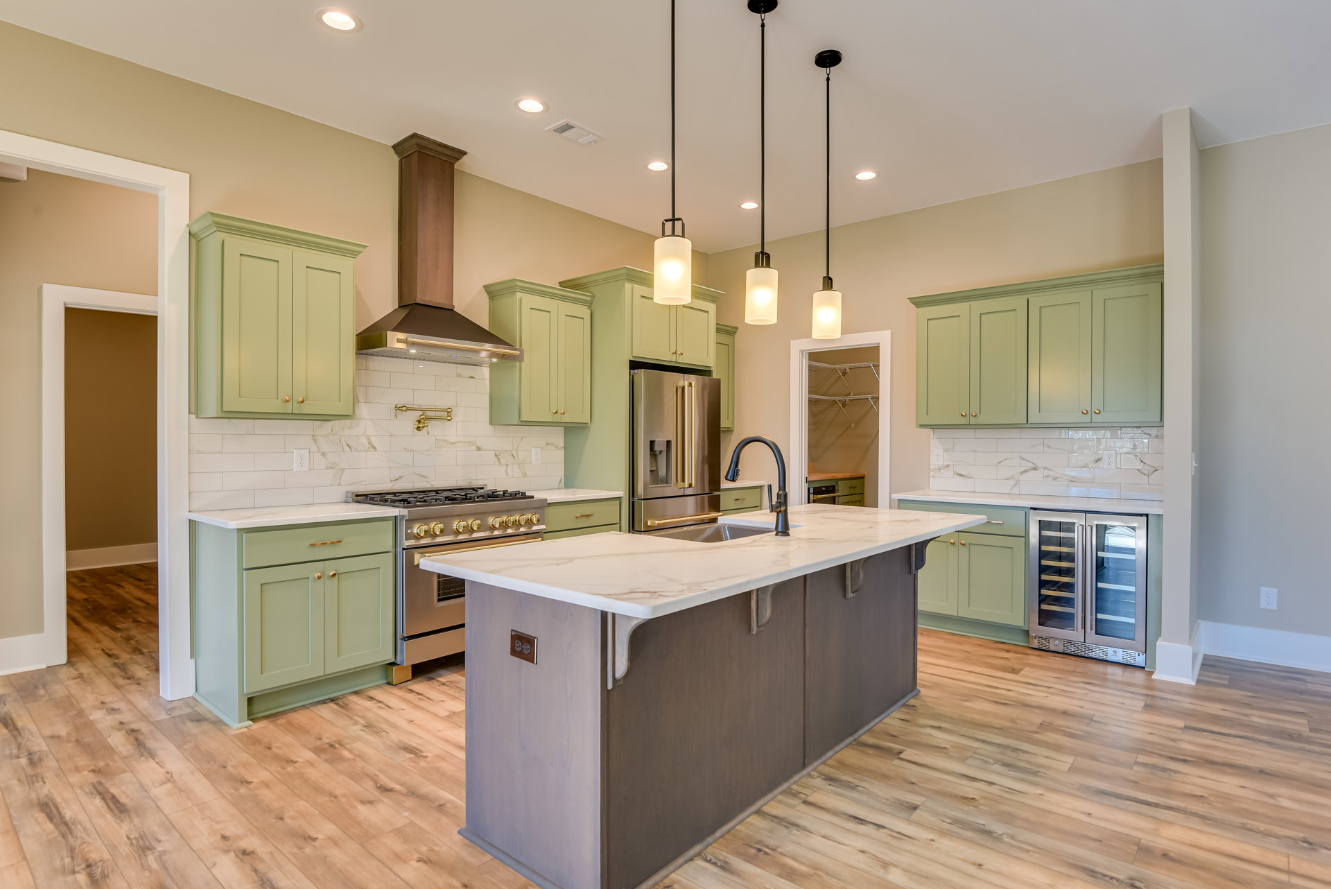 Green kitchen cabinets with brass hardware, large central island featuring a built-in sink and white quartz countertop, stainless steel refrigerator, pendant light fixtures above