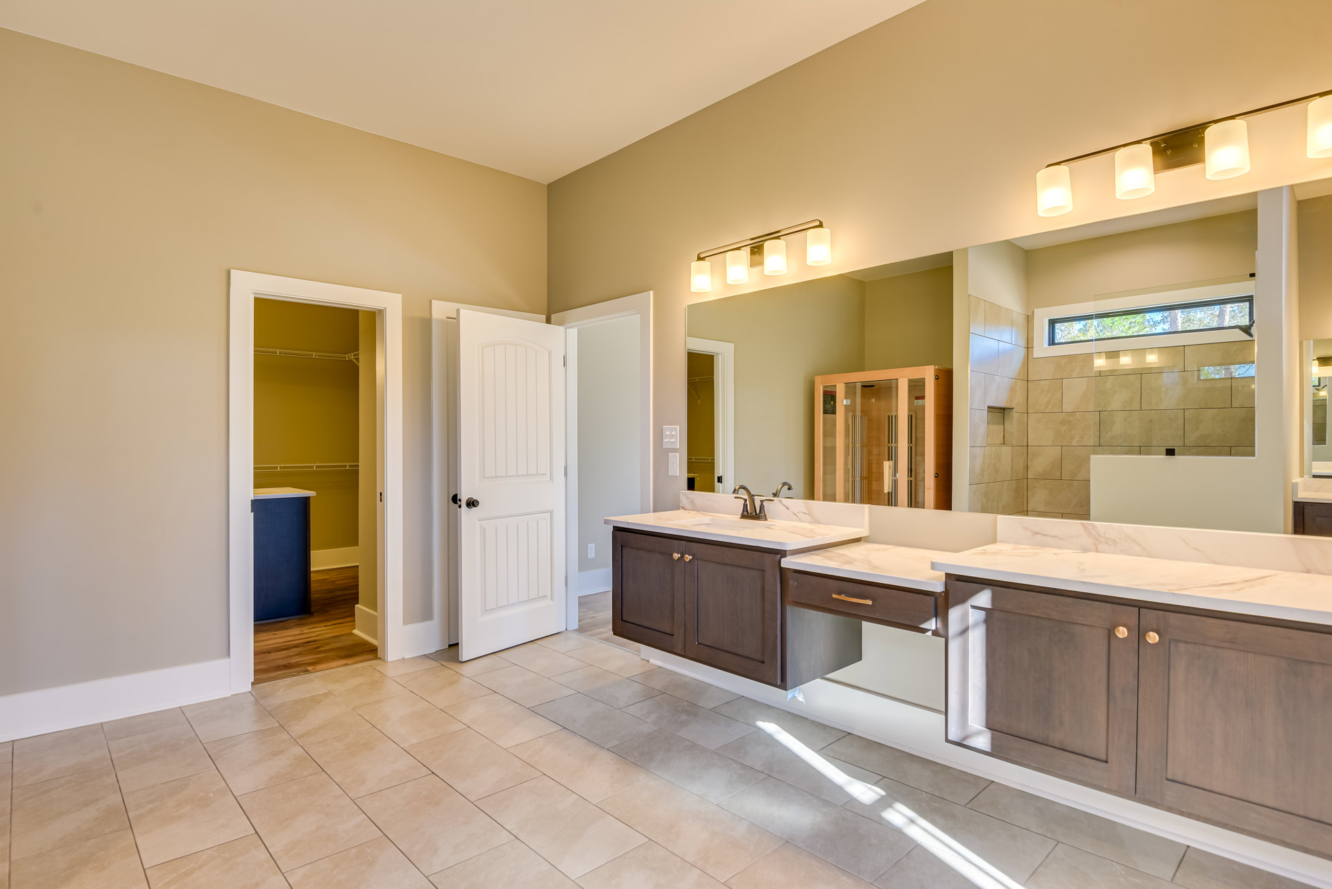 Bathroom with double sinks set in a light stone countertop, large frameless mirror above, white door with black knob, wooden cabinet with glass doors, closet with white frame