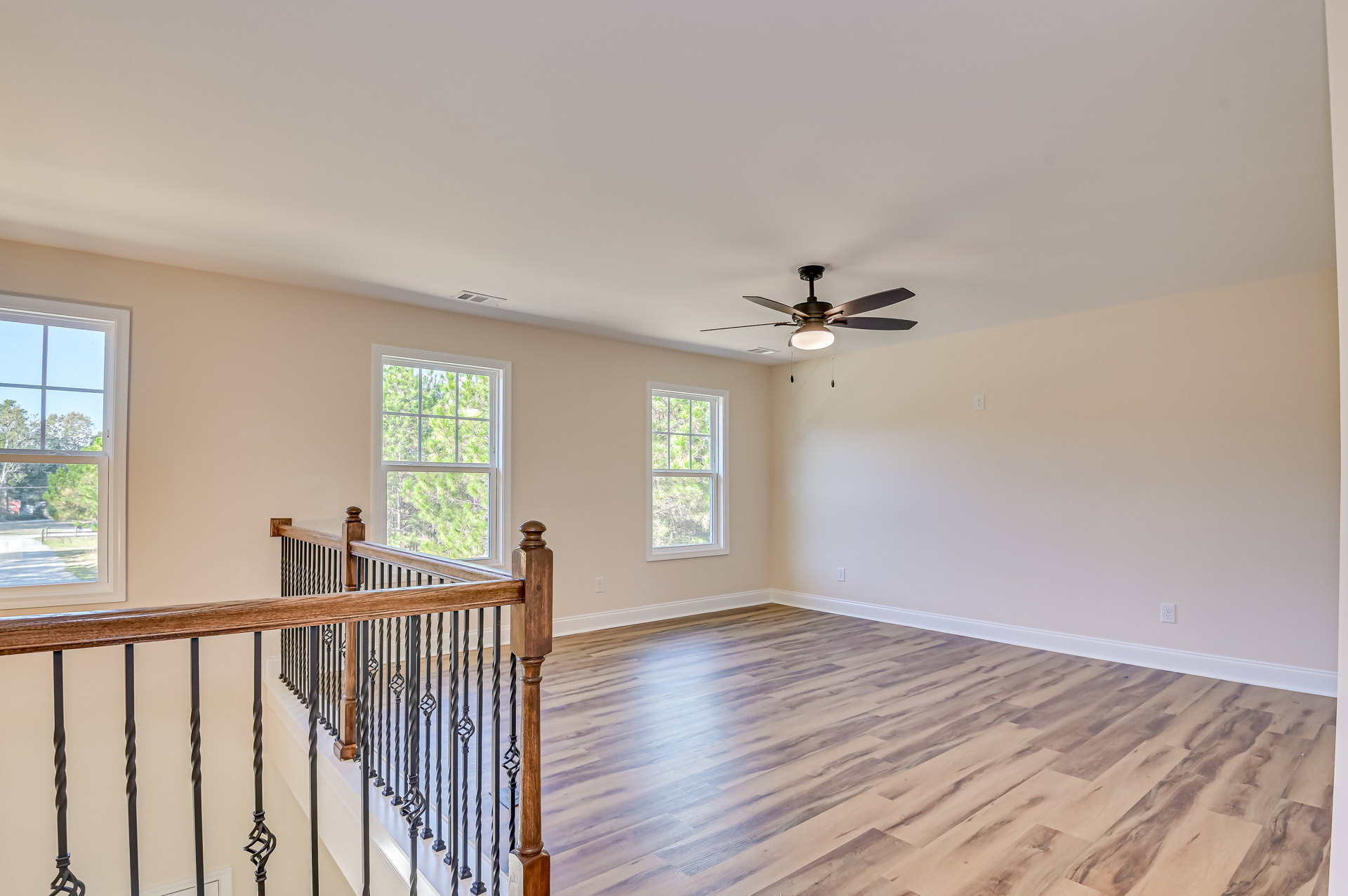 Wood floor room with white walls, ceiling fan with light, wood staircase railing, large window overlooking trees