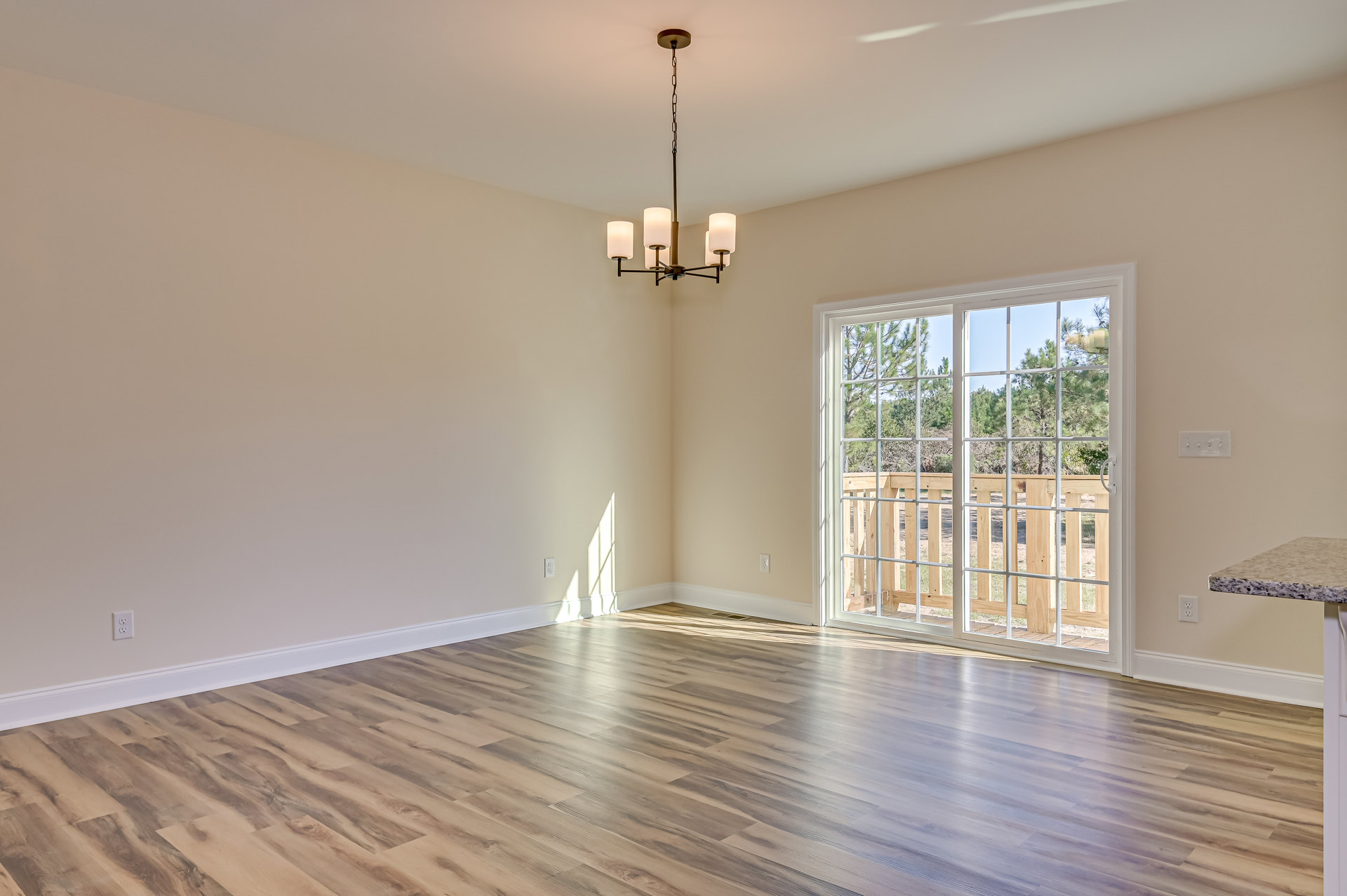 Sunlit hardwood floor in a room with white plaster walls, large window framed by wood railing, modern ceiling light fixture suspended by chain, trees visible outside