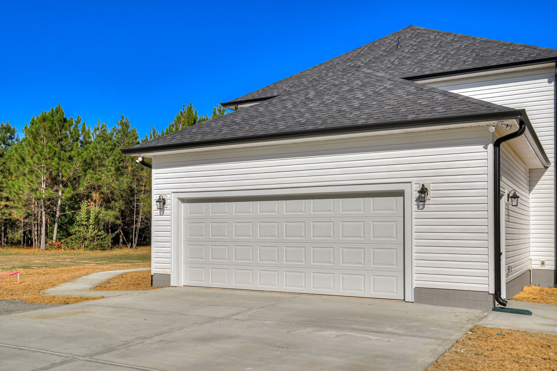White garage door beneath black roof, concrete driveway, mature trees in background, light post near fence with red tape, white rectangular object with square border on exterior