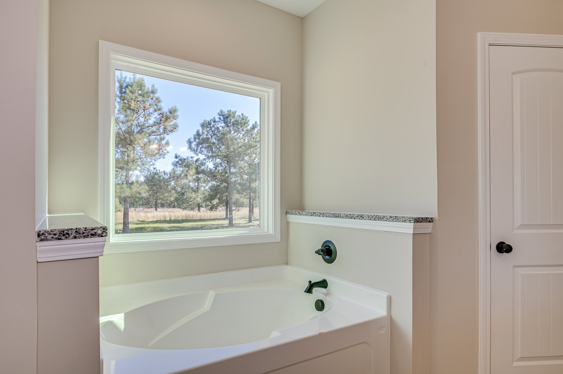 White freestanding bathtub beneath a large window with leafy trees visible outside, black faucet mounted on white tile, white door with black handle, minimal bathroom decor.