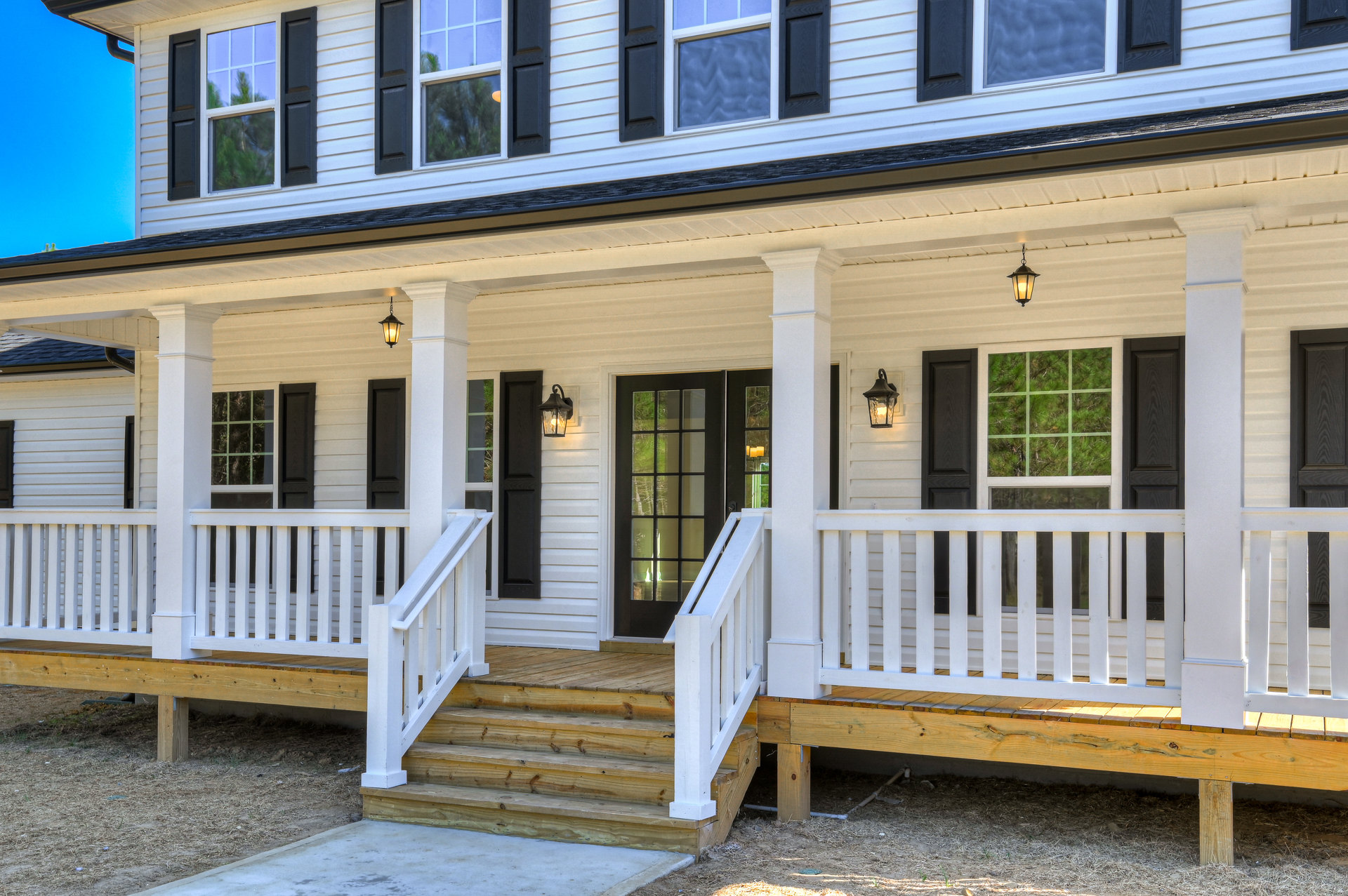 White house exterior with black shutters, wooden porch and stairs, glass-paneled front door, white railings, and blue sky background