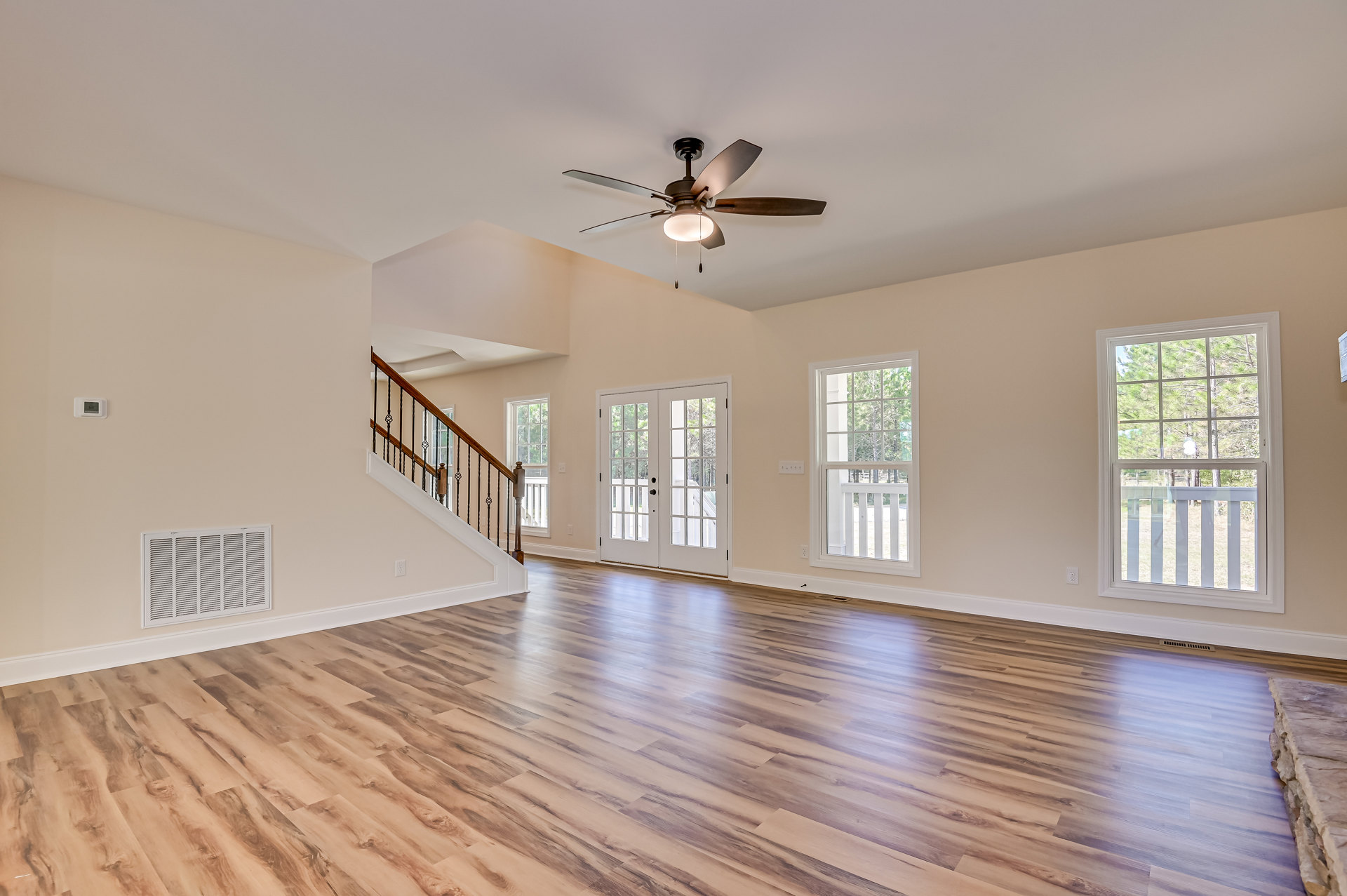 Spacious interior with hardwood floors, white walls, ceiling fan with light, double glass-paneled doors, wood-railed staircase, and window overlooking trees