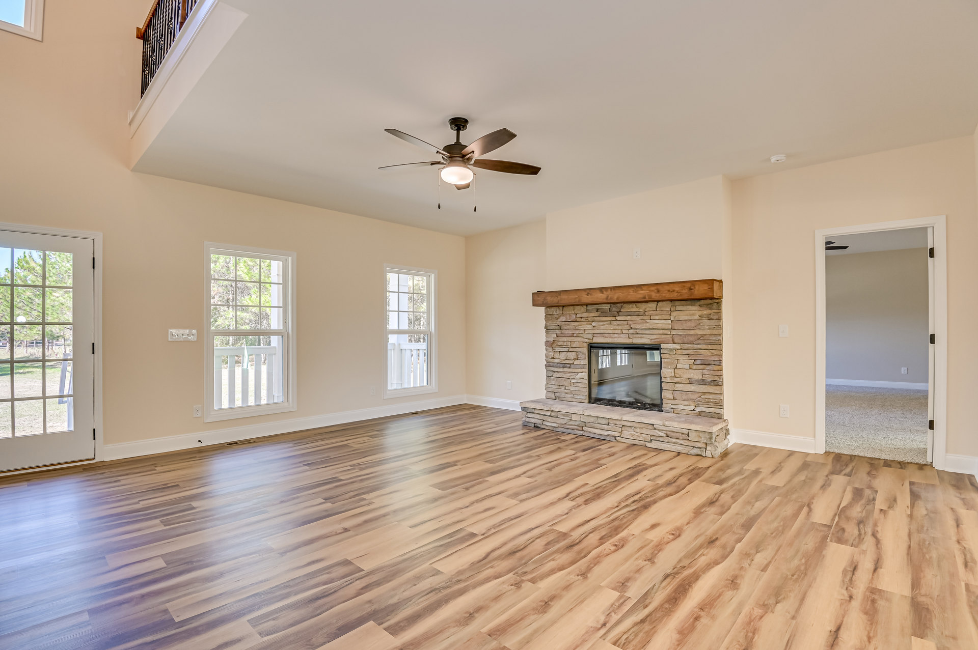 Living room with hardwood floors, brick accent wall featuring a glass window, ceiling fan with light fixture, fireplace topped by a wood beam, windows overlooking yard and trees
