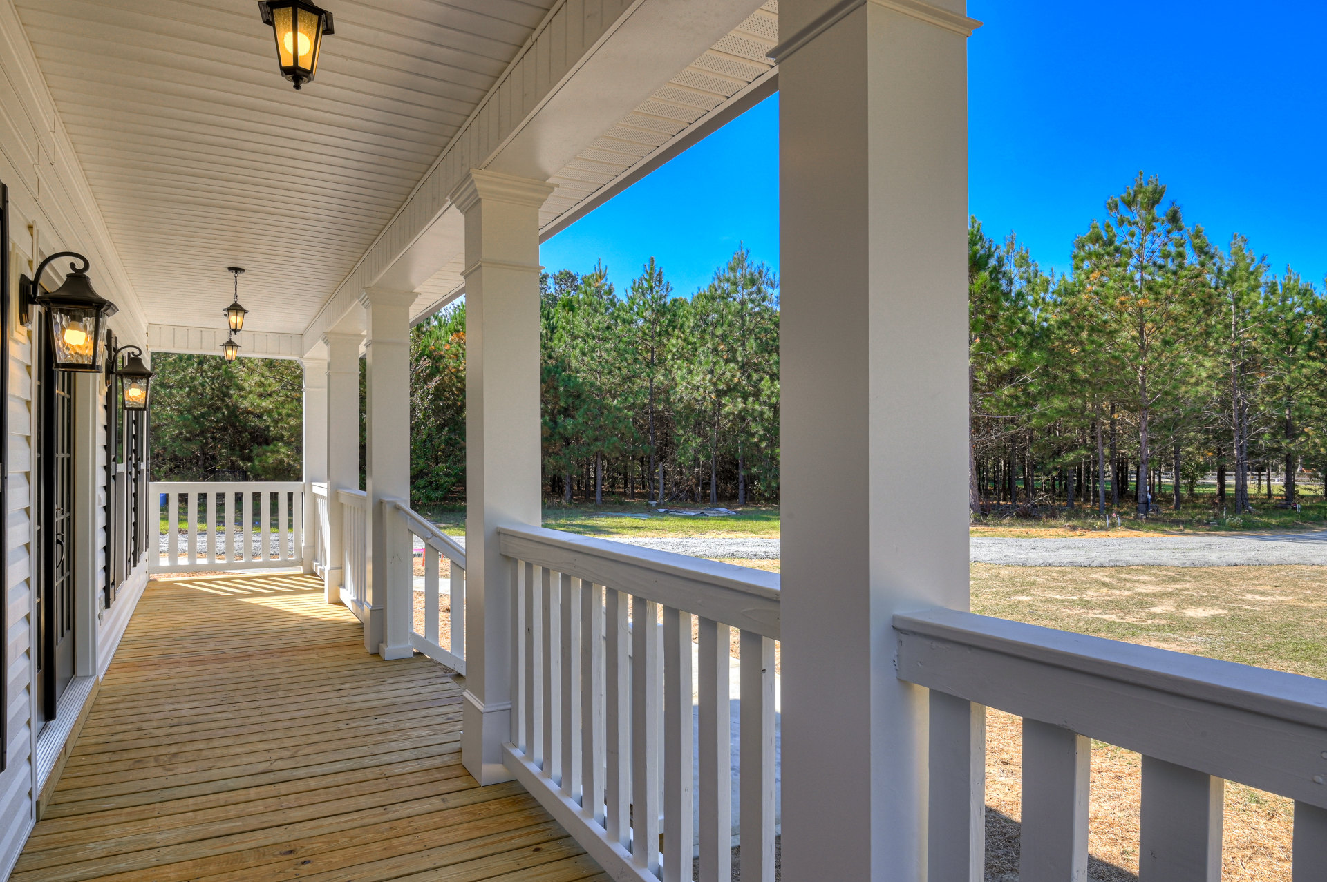 Wide porch with white pillars, wood floor, white railings, lit ceiling fixture, and a white fence bordering a tree-lined path in the background