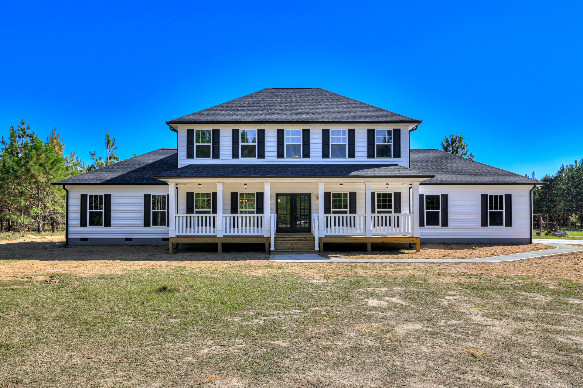 White two-story house with wide front porch, upper balcony, double glass-paneled doors, multiple windows, wooden steps, and manicured green lawn bordered by trees.