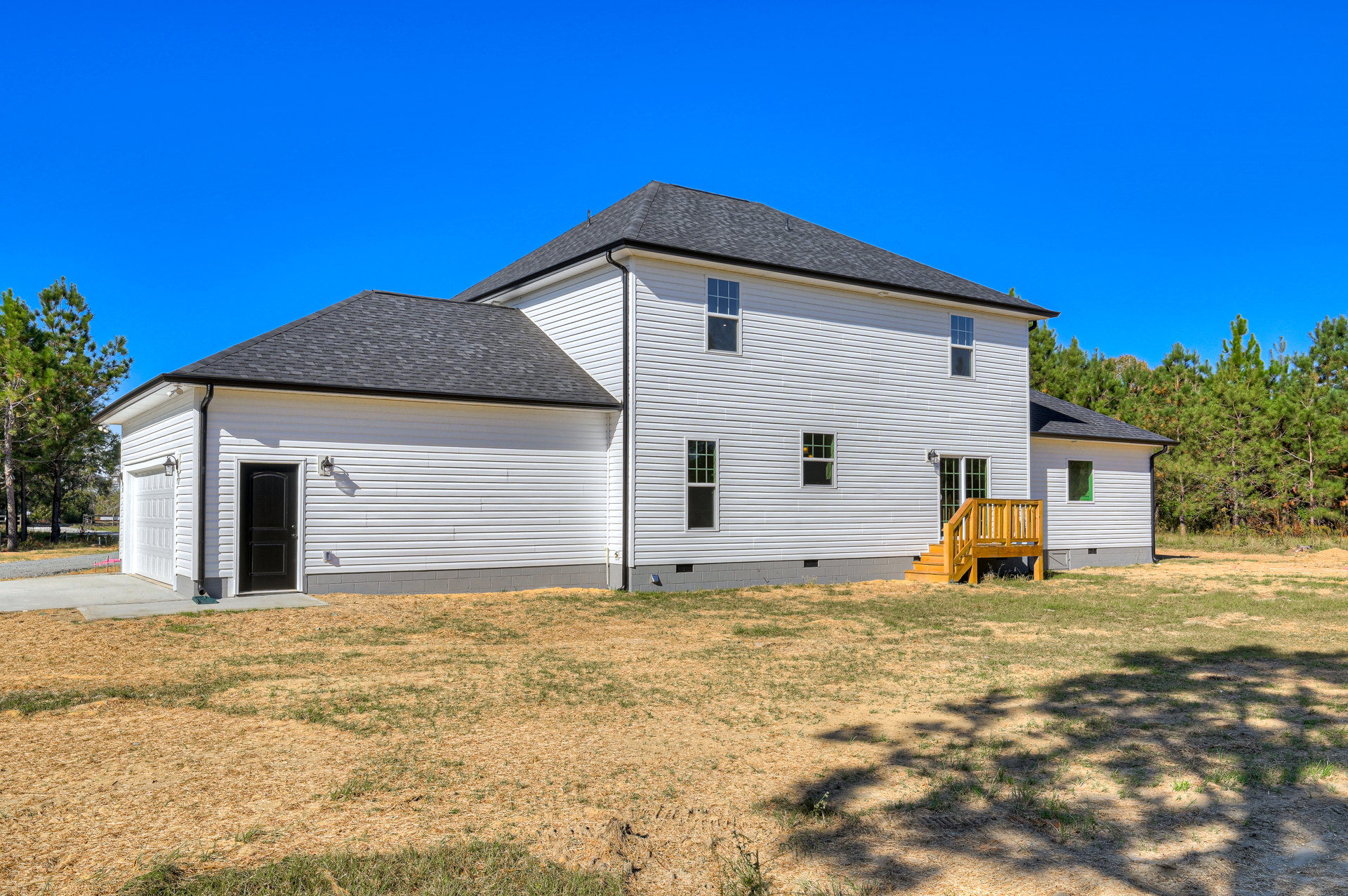 White siding house with black roof and black front door, wooden deck with stairs leading to grassy yard, mature trees and tree shadows visible in background.
