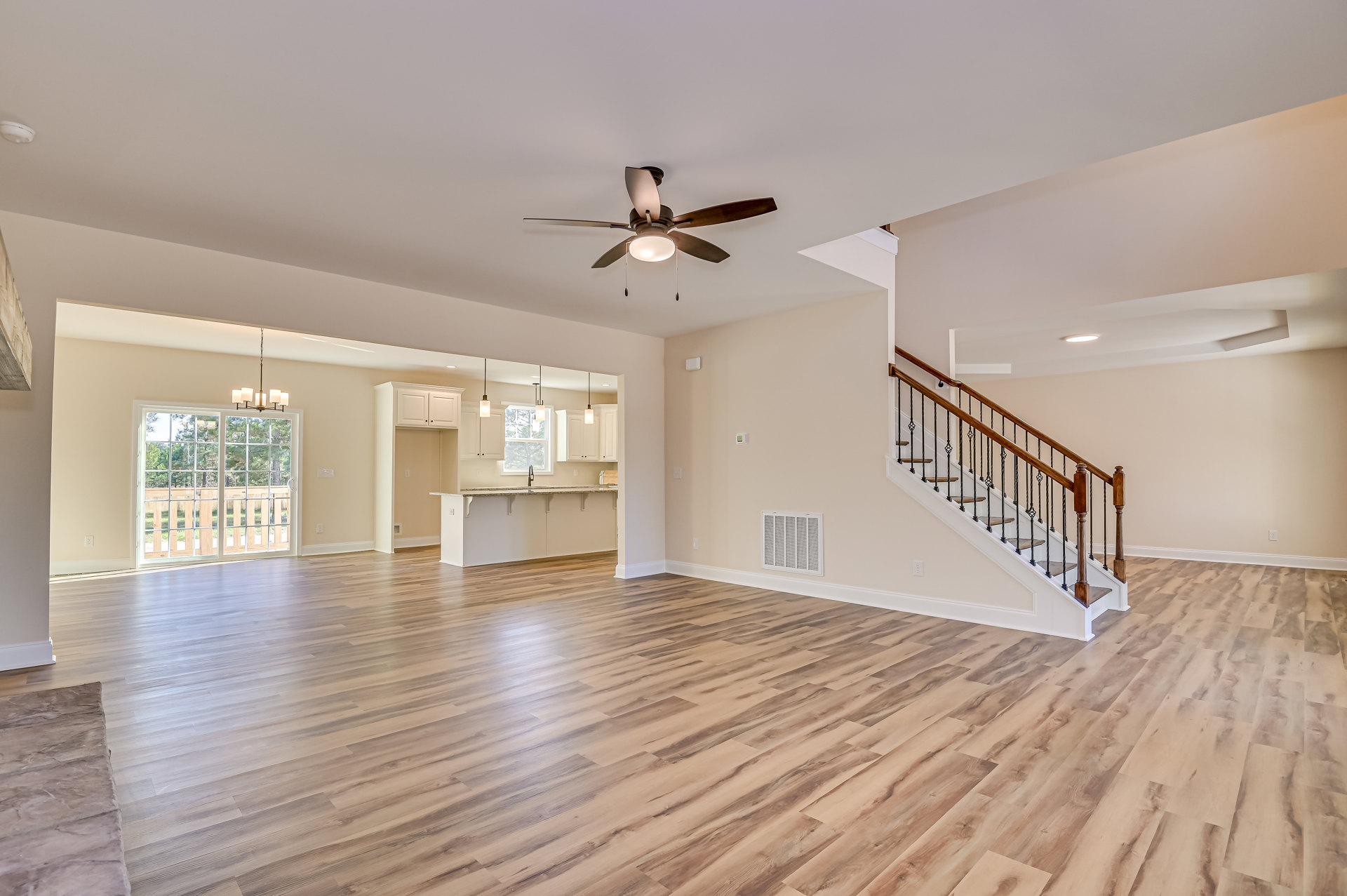 Spacious living area with hardwood flooring, ceiling fan with light, wood staircase railing, window showing trees outside, and wall vent