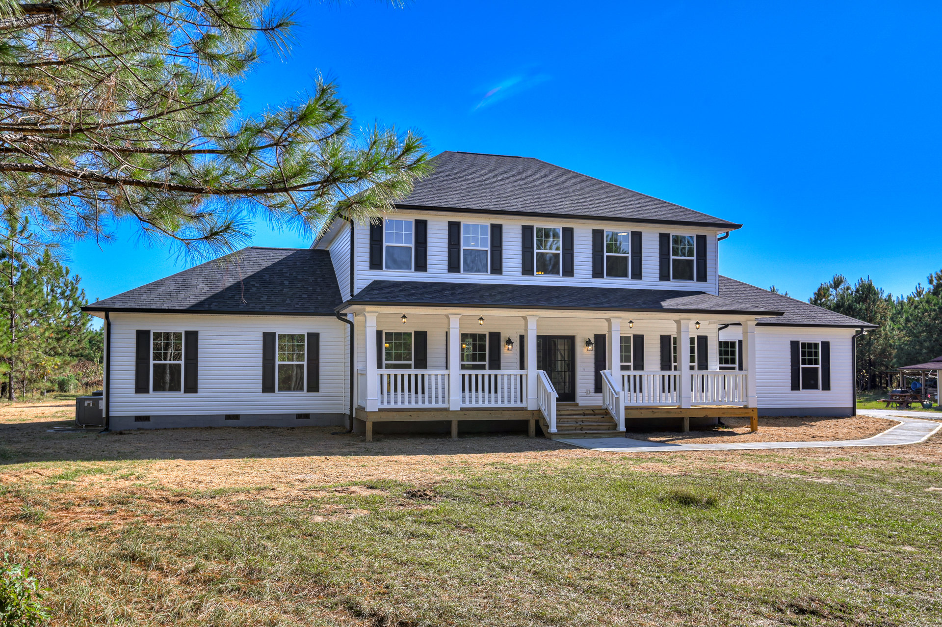 White two-story house with wide front porch, white railings, large windows, manicured green lawn, mature tree, and blue sky overhead